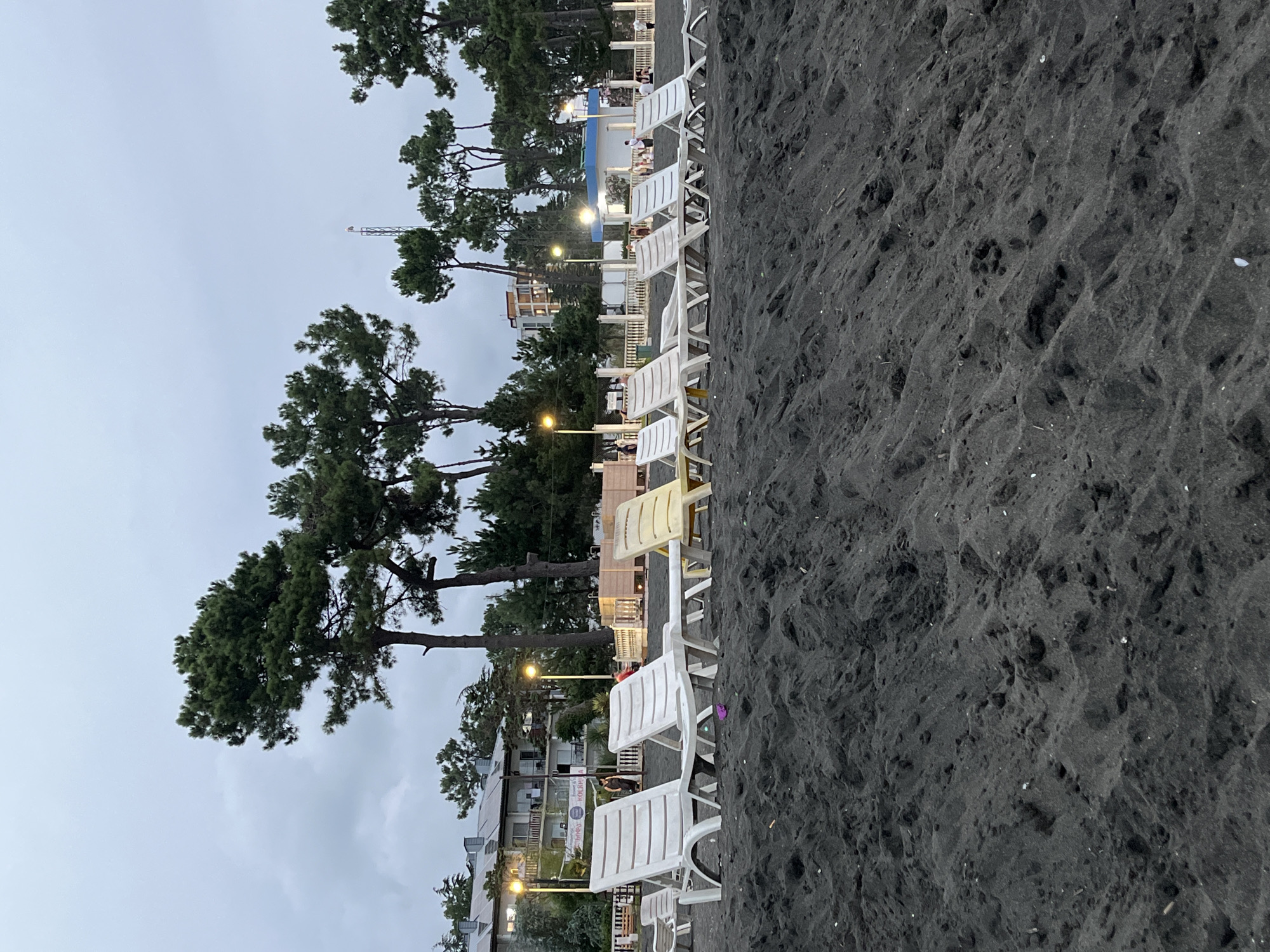 The image depicts a beach scene in Ureki, Georgia, characterized by dark volcanic sand. In the foreground, the sand is piled high, showing textured ripples and tracks. A row of white plastic lounge chairs is arranged on the sand, with a single yellow lounge chair among them. In the background, several tall pine trees stand against an overcast sky. Behind the trees, a building with balconies and lights is visible, suggesting a hotel or resort. A white fence separates the beach from the building and surrounding greenery. Several people are present in the background, some standing and others appearing to be seated or walking along the beach. The overall atmosphere is calm and appears to be twilight or an overcast day.