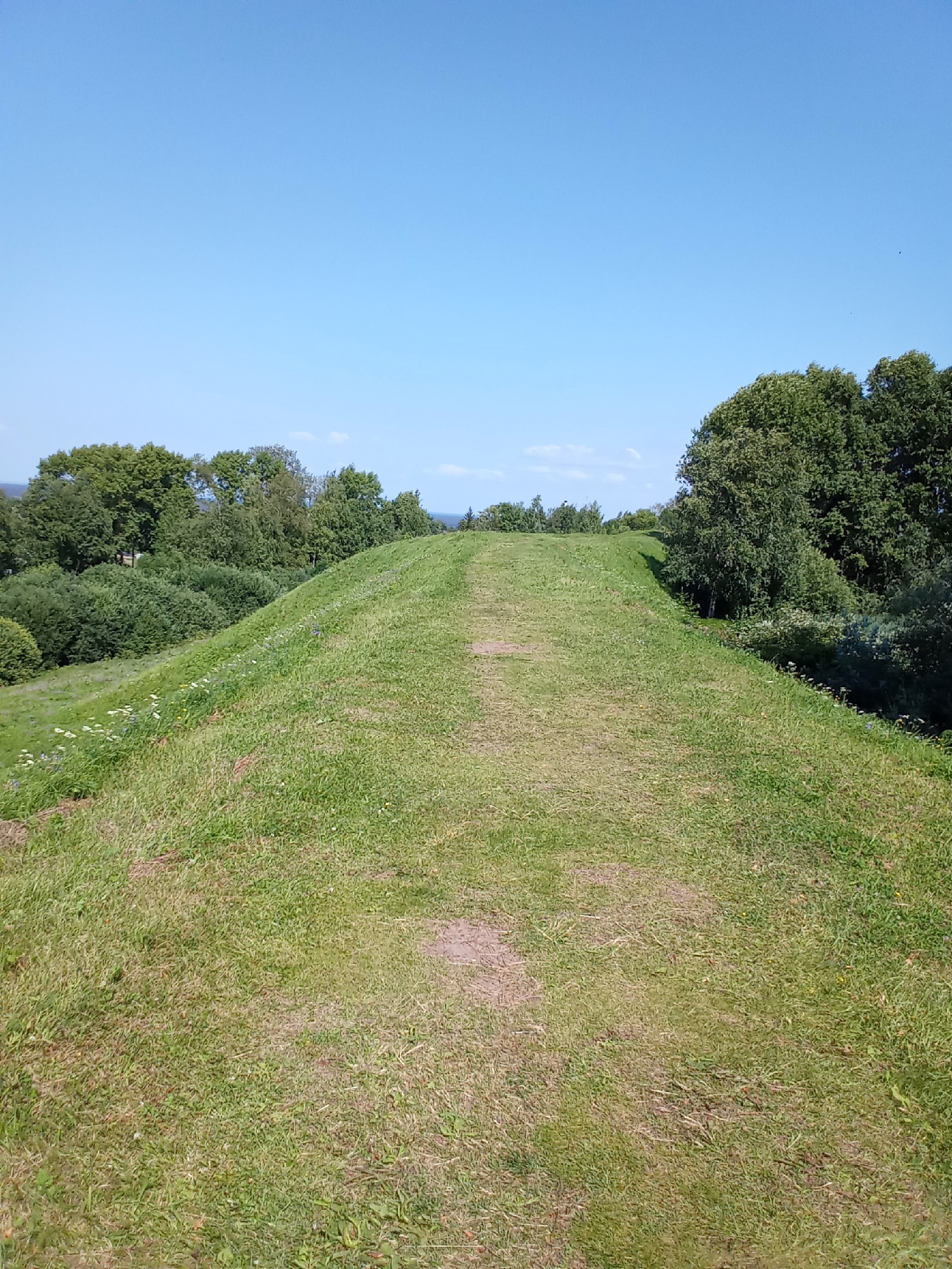 The image shows a wide, grassy hill with a path running up its center. The hill is covered in green grass with scattered patches of bare earth. In the background, there are dense trees and a bright blue sky with a few wispy clouds. The scene appears to be a natural landscape, possibly a park or a preserved historical site. Based on its appearance, this could be the Belozersky Kremlin, a historical complex located in the city of Belozersk, Russia, known for its large defensive earthworks.