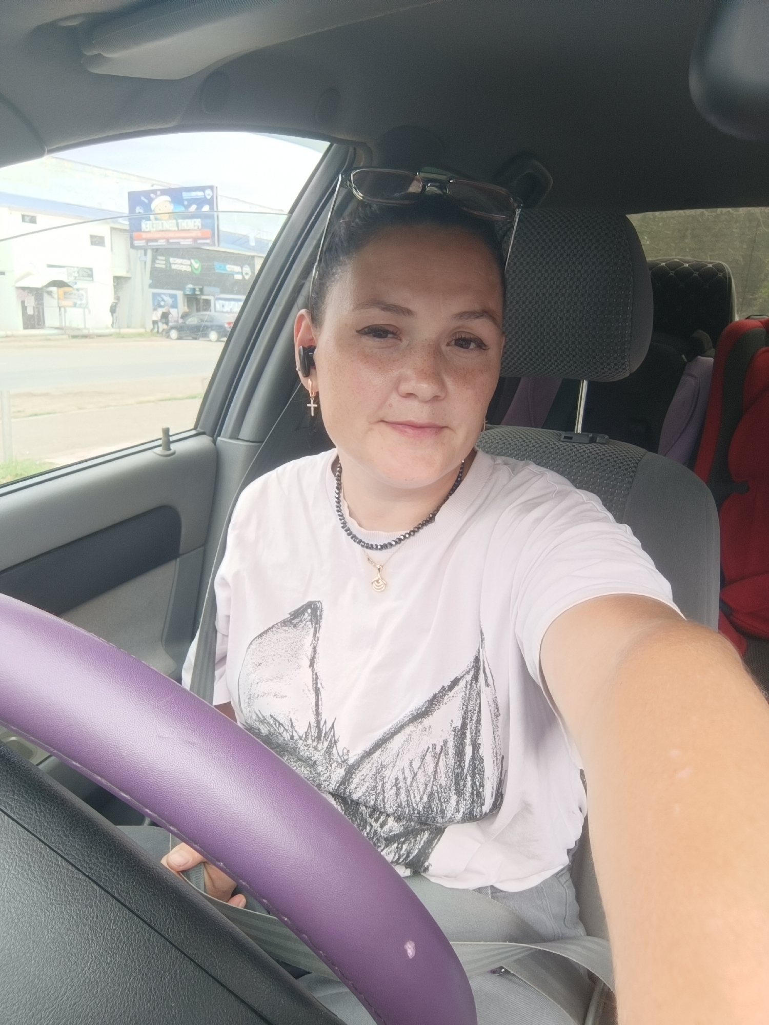 A woman with freckles, wearing a white t-shirt, black earbuds, and a necklace with a cross pendant, is sitting in the driver's seat of a car. She is wearing glasses on top of her head. A purple steering wheel cover is visible in the foreground. The background shows buildings and a billboard in the distance, suggesting an urban environment. The scene appears to be in the city of Chaikovsky, Russia.