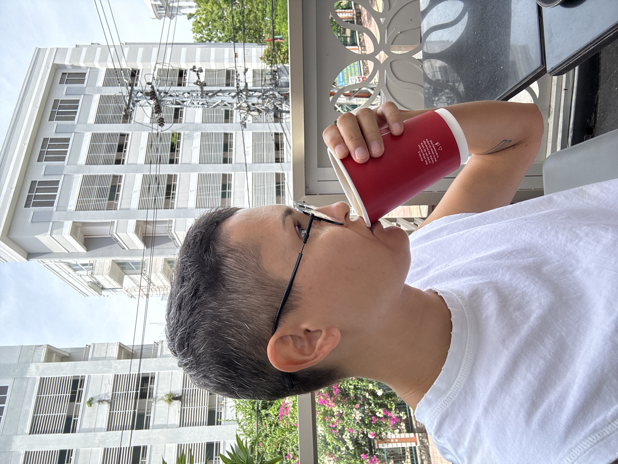 An individual with short, dark hair and glasses is seen in profile, drinking from a red paper cup. They are wearing a white t-shirt. The background features two tall, grey buildings with many horizontal windows, and a utility pole with electrical wires. To the left of the frame, there is a bush with pink and white flowers. The scene appears to be outdoors, possibly on a balcony or patio, indicated by a table in the foreground and a decorative railing. The buildings and utility pole suggest an urban environment.