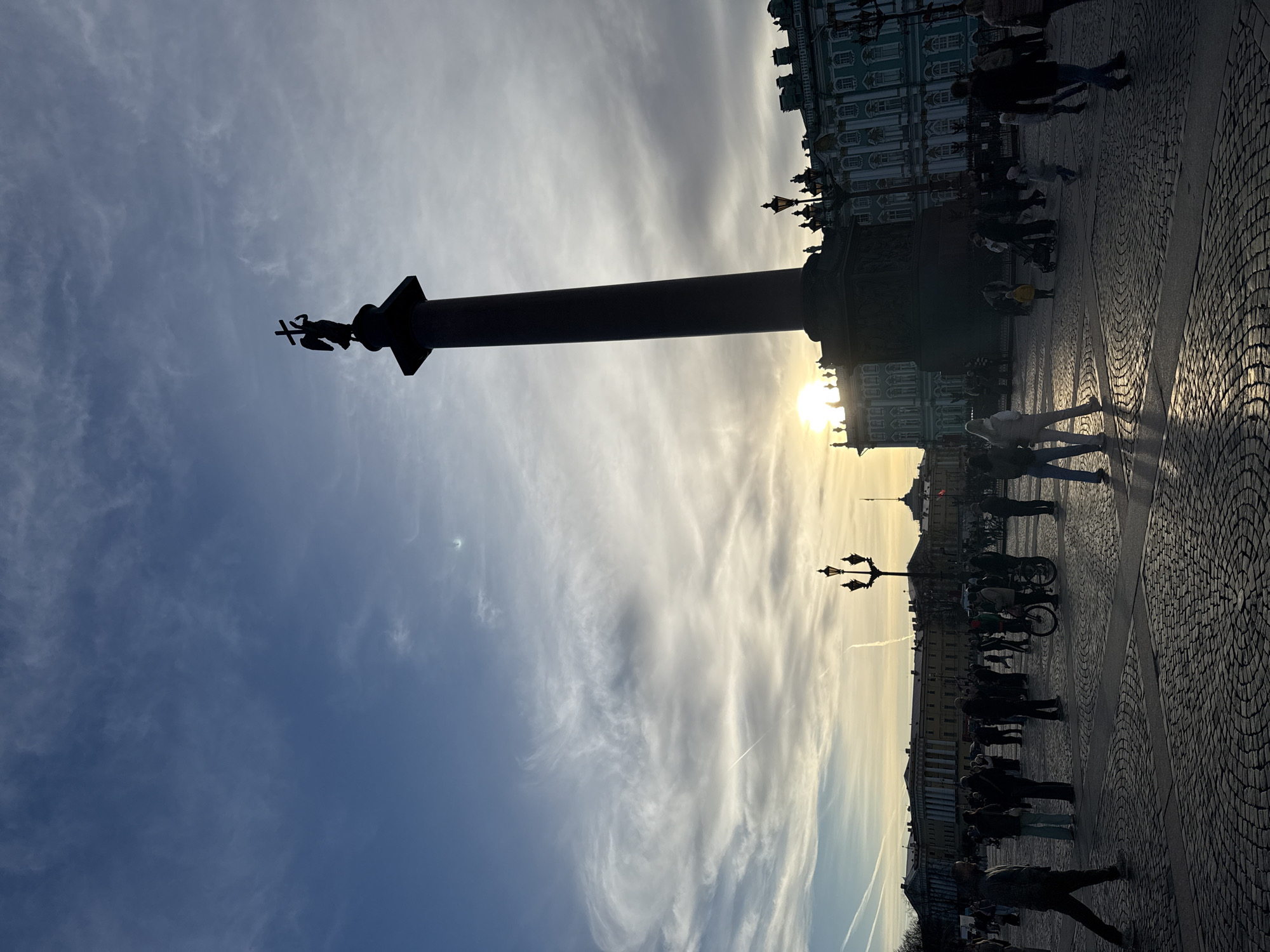 The image captures a vibrant public square in Saint Petersburg, Russia, bathed in the dramatic light of either sunrise or sunset. The central and most prominent feature is the colossal Alexander Column, which stands tall, its dark silhouette dominating the left side of the frame. Perched atop the column, a statue of an angel holding a cross is clearly visible, also in silhouette against the bright sky.
To the right of the column, the stately Winter Palace (Hermitage Museum) extends across the midground. Its ornate architecture and distinctive color are somewhat obscured by the backlight, yet its grandeur is evident. Further into the distance on the right, additional buildings line the horizon.
The foreground and midground of the square are covered in cobblestones, with areas illuminated by the low-angled sunlight creating a textured pattern of light and shadow. Numerous people are present, their forms rendered in silhouette against the bright ground and sky. They appear to be strolling, standing, and gathering, suggesting a typical day in this popular public space. A distinctive street lamp, also silhouetted, is visible to the right of the column, and hints of bicycles can be seen among the crowd.
The sky is a significant element, transitioning from a deep blue on the far left to a brilliant, warm yellow and orange glow where the sun dips behind or emerges from the horizon. Wispy, streaky clouds stretch across the sky, catching the golden light and adding to the dramatic atmosphere. The strong backlighting emphasizes the silhouettes of the column, people, and architectural details, creating a striking contrast between light and shadow. No visible text is present in the image.