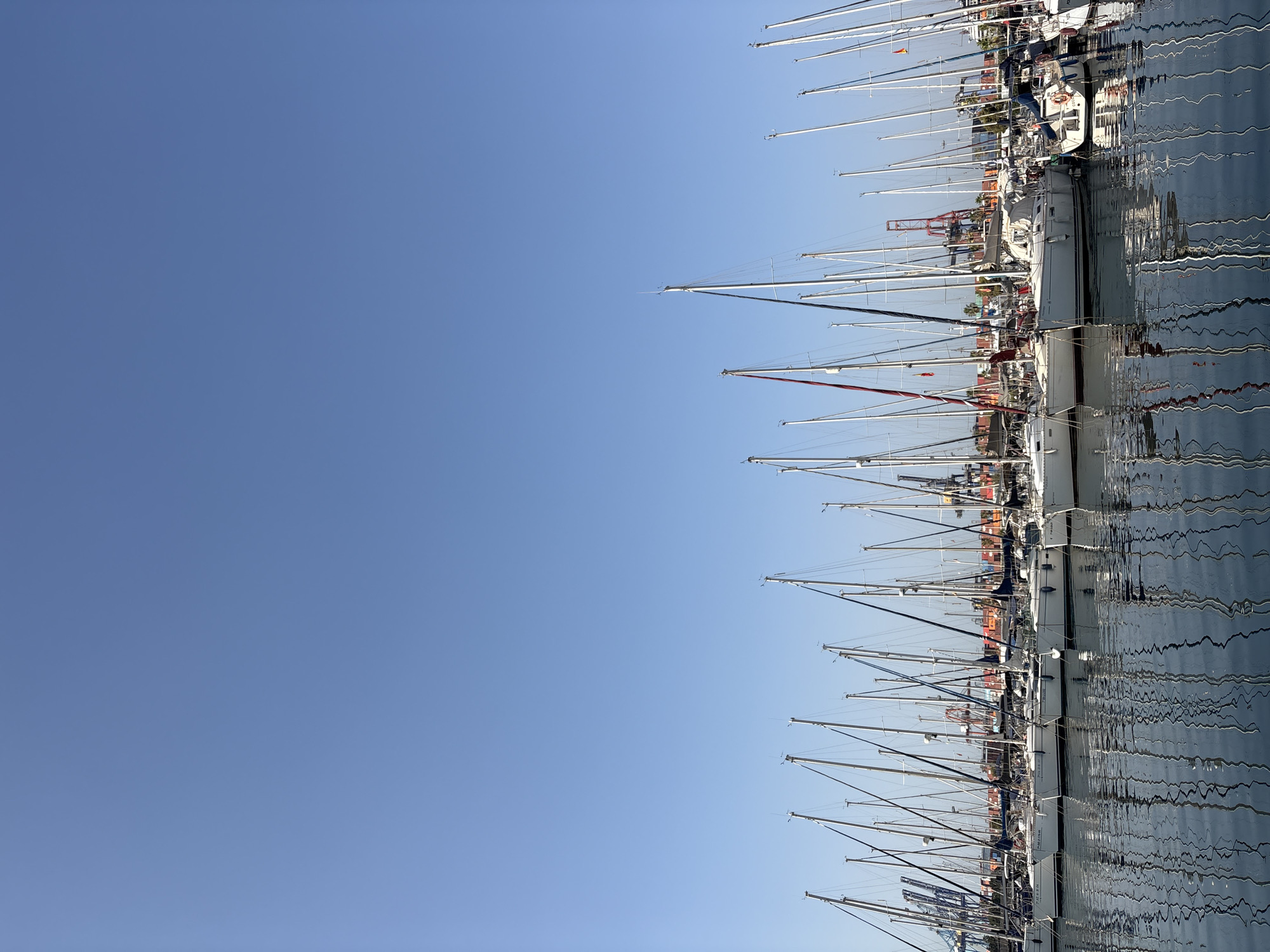 A wide-angle, eye-level shot depicts a marina in the city of Valencia, Spain, under a clear blue sky. The foreground is dominated by the calm, reflective surface of the water, mirroring the numerous sailboats docked in the marina. These sailboats are white with tall masts, many of which are rigged with sails that are furled. They are lined up neatly, suggesting organized mooring. In the background, beyond the sailboats, a commercial port is visible with large cranes and shipping containers, indicating a bustling industrial area adjacent to the recreational marina. The horizon line is low in the frame, with the majority of the image dedicated to the expanse of the sky. There are no people visible in the frame. The overall scene is one of a serene marina set against a backdrop of industrial activity.