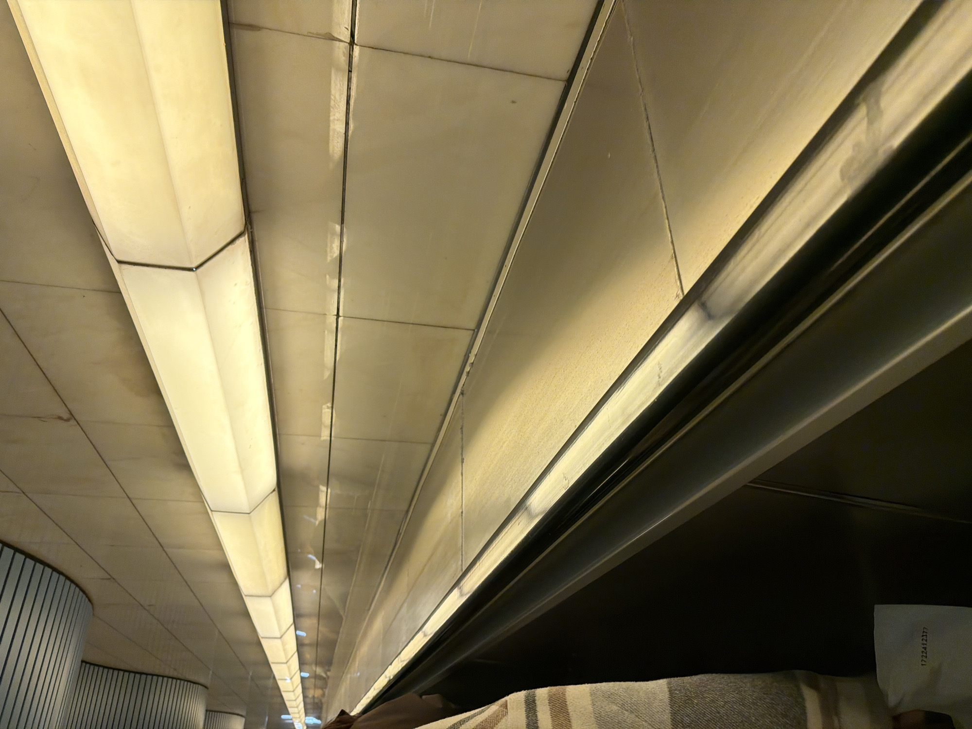 The image depicts the interior of a night train compartment in Moscow, Russia. The perspective is from a bottom bunk looking upwards. The ceiling is composed of rectangular tiles with linear light fixtures running along their edges. The upper part of the bunk structure is visible, made of metal and dark-colored material. A striped blanket is partially visible on the lower bunk. In the lower right corner, a partially visible paper with numerical markings is present. The light fixtures illuminate the ceiling. Vertical striped panels are visible in the background.