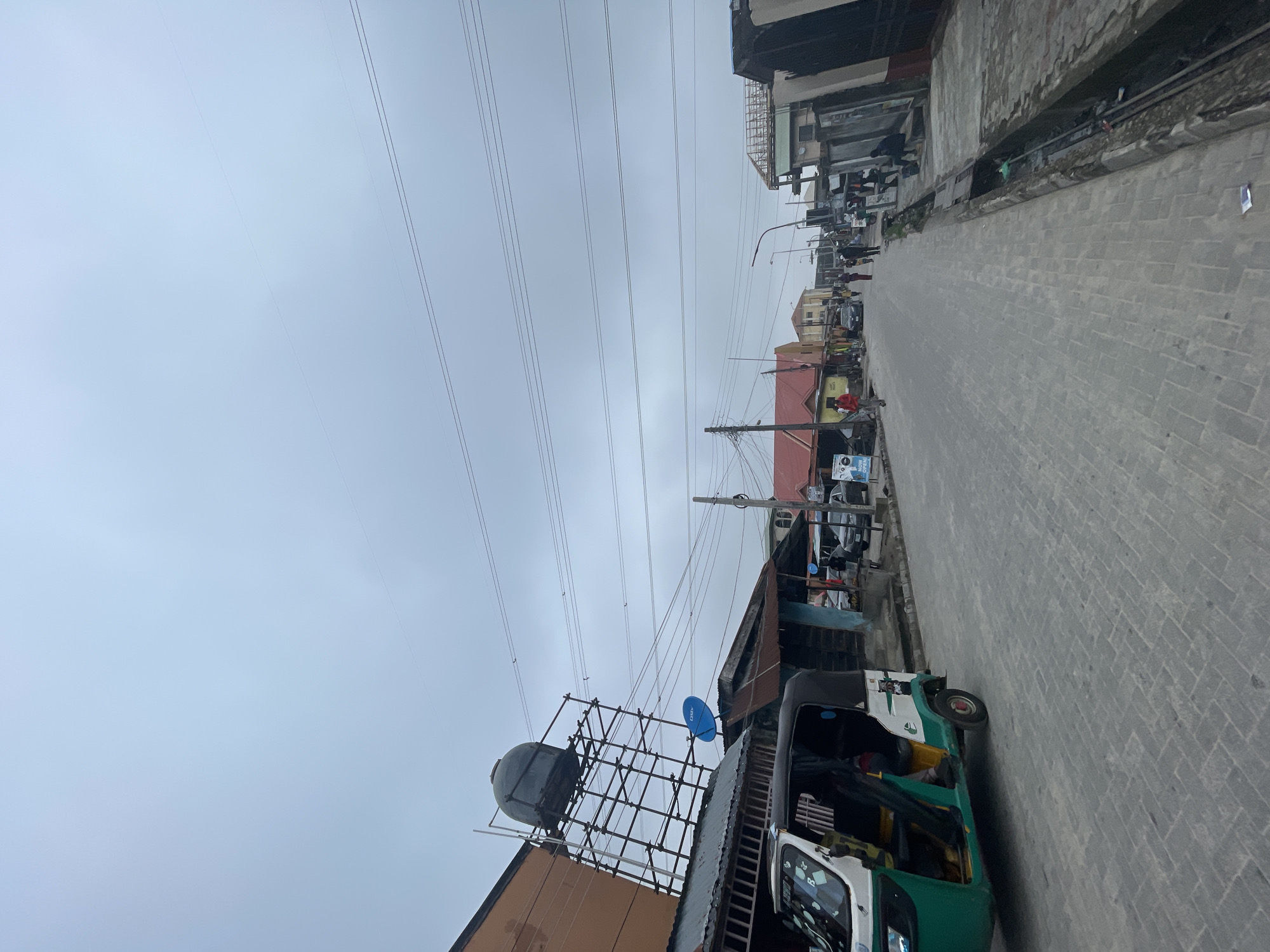 The scene depicts an urban street in Ado, Nigeria, under a uniformly overcast sky. The road is paved with grey interlocking pavers, extending into the distant vanishing point.
On the immediate left, a green and white three-wheeled motorized rickshaw (keke napep) is partially visible. Behind it, a multi-story brown building supports a large, dark, spherical water tank elevated on a robust metal scaffolding structure. Below the water tank, a building with a corrugated metal roof features a blue circular satellite dish. Further down the left side, a building with a distinct red gable roof is visible, in front of which a white sign displaying "NOW OPEN!" in blue text is present. Multiple individuals are walking along the left side of the street, including one person in a red top near the "NOW OPEN!" sign.
On the right side of the road, an open concrete drainage channel runs parallel to the pavement, containing minor debris. Various buildings with differing architectural styles and states of repair, including one with exposed roof rafters, line this side of the street. Several pedestrians are observed walking along the right sidewalk and road, receding into the background. One individual is seated on a low concrete structure on the right sidewalk in the mid-ground.
Overhead, multiple dark utility wires span horizontally across the upper half of the image, supported by several vertical utility poles. A street light fixture extends over the road in the mid-distance. Additional vehicles, including cars, are discernible further down the street on both sides.