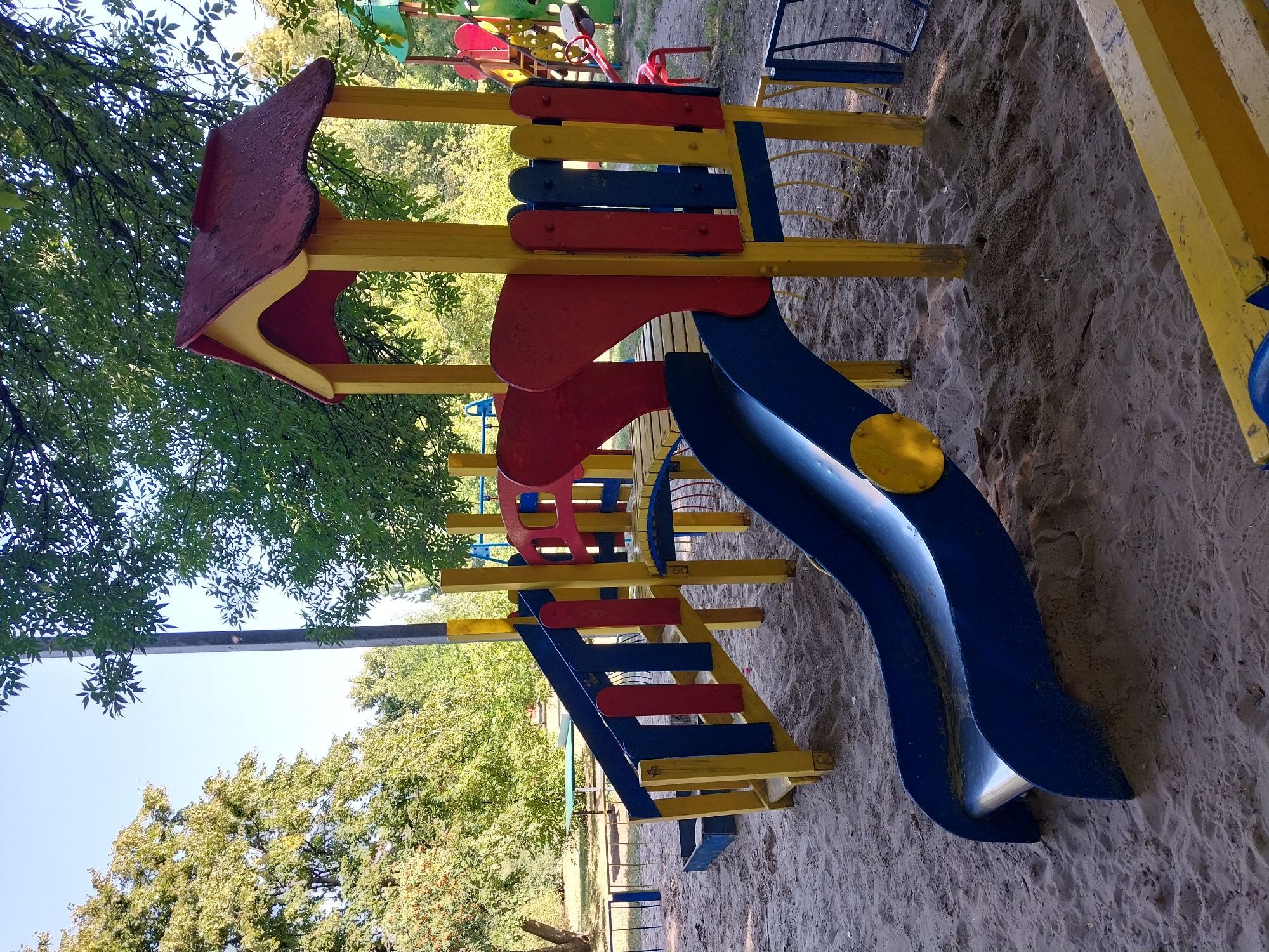 The image shows a colorful playground with slides and climbing structures made of yellow, blue, and red painted wood. The playground is situated on a sandy surface, with trees and foliage in the background. In the distance, a green picnic table can be seen. The scene appears to be set on a clear day with sunlight illuminating the area. There are no people visible in the image. The location is identified as Doslidne, Ukraine.
