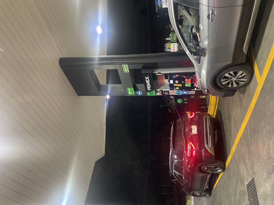 A gas station at night with two vehicles. A silver SUV is pulled up to a PEMEX pump, appearing to be refueling. Behind it, a dark-colored SUV sits, its taillights illuminated, potentially waiting to fuel. The pump displays various buttons and displays, including a "10 PEMEX" label. Yellow lines mark parking spaces on the concrete surface. The background is dark, with faint lighting from nearby structures.