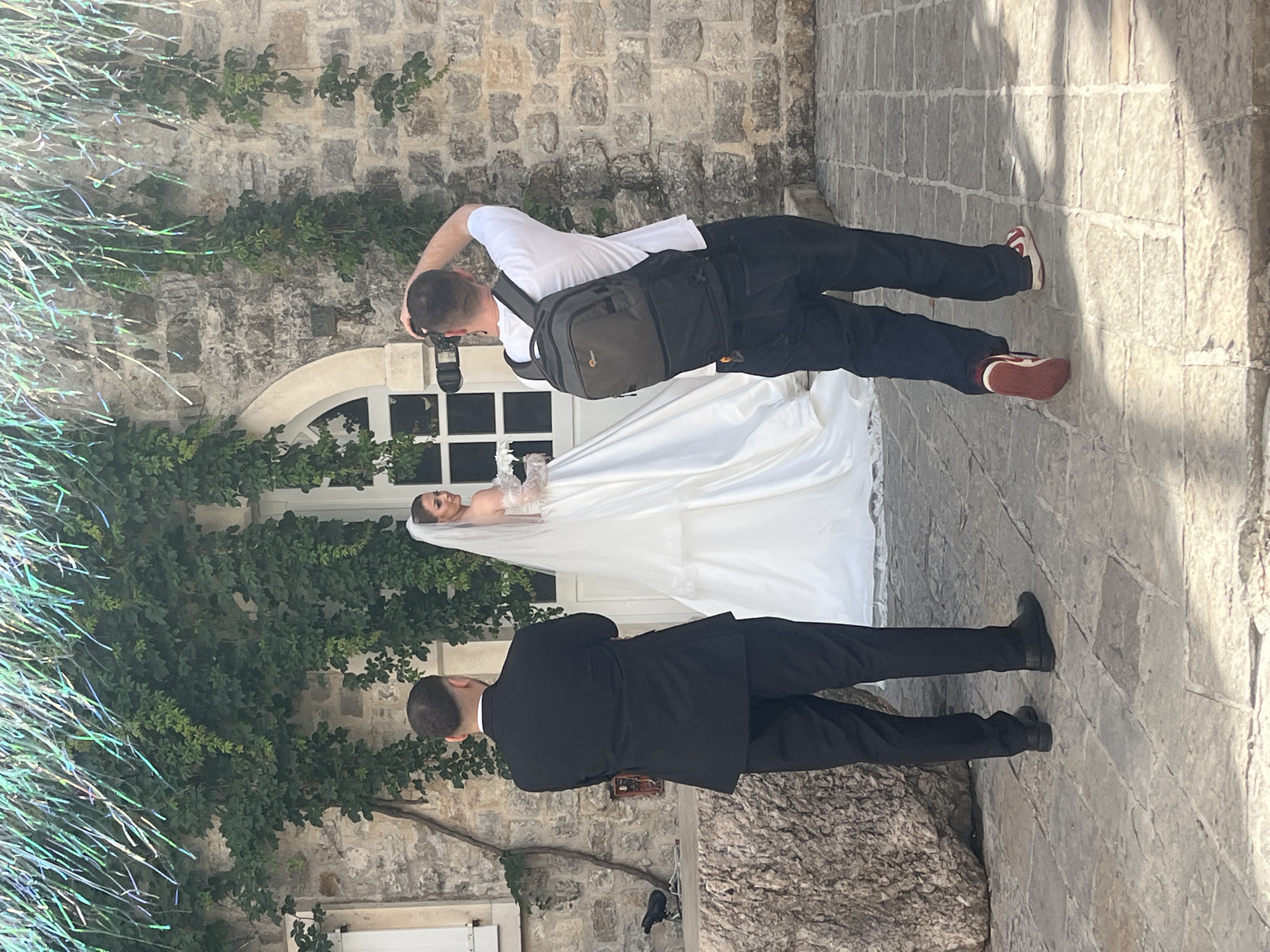 The image captures an outdoor wedding photoshoot featuring a bride, a groom, and a photographer, set against a historic stone wall backdrop in what appears to be Budva, Montenegro.
In the center, a male photographer, viewed from behind, is focused on taking a picture of the bride. He wears a white t-shirt, black trousers, and red and white sneakers. A dark grey camera bag is slung over his right shoulder, and he holds a black DSLR camera equipped with a flash.
The bride, positioned to the photographer's left, is wearing a white wedding gown and a veil. She is seen from the chest up, posing in front of a white arched window embedded in the stone wall. The wall itself is covered with patches of green ivy.
To the far left, a groom, also seen from behind, stands observing the scene. He is dressed in a formal black suit and black shoes. The left side of the frame is adorned with shimmering, tinsel-like decorative streamers, possibly indicating a festive event or a prop.
The ground is paved with flagstones, adding to the rustic and historic ambiance. The scene is brightly lit, suggesting it is daytime. The activity is clearly a professional wedding photography session in a scenic, old-world location. No explicit text is visible in the image.
