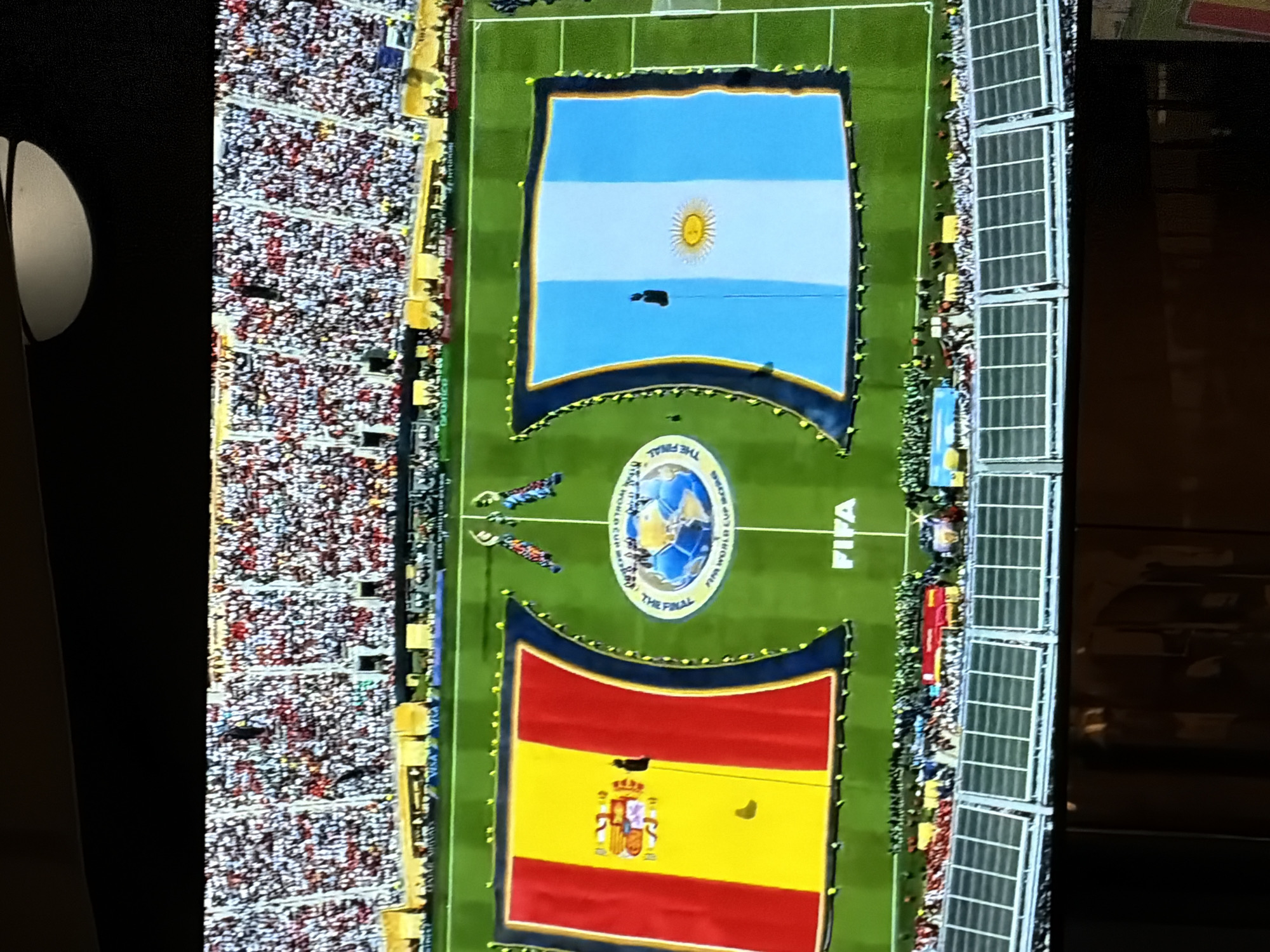 The image captures a wide aerial view of a soccer stadium during an event, likely a pre-match ceremony. The field is largely covered by two massive flags, one representing Spain and the other Argentina. These flags are unfurled across the pitch, with a FIFA World Cup logo in the center, suggesting a significant international match.
On the left, the Spanish flag, featuring red and yellow stripes with a coat of arms, is prominently displayed. To its right, the Argentine flag, with horizontal light blue and white stripes and a sun emblem, is equally visible. Both flags are held in place by numerous people positioned around their edges, forming a human border.
The stadium seating, packed with a vast crowd, encircles the field. The spectators are a blur of colors, predominantly white and red, indicating fan support for one or both teams. In the background, the stadium architecture is visible, including tiered seating and possibly advertising boards.
The foreground shows the lower levels of the stadium, including what appear to be concourse areas and further seating. The overall atmosphere suggests a grand sporting occasion, with a significant display of national pride and fan engagement. The presence of the FIFA World Cup logo confirms this as a high-profile soccer event.
The provided location of Budva, Montenegro, is not visually supported by any identifiable landmarks or context within the image. The scene is generic to a large soccer stadium and does not contain any elements that would place it specifically in Budva.