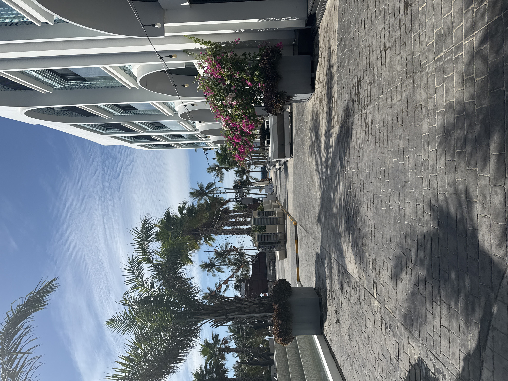 A paved walkway with a stamped brick pattern leads alongside a white, modern building with arched windows and walkways. Palm trees line the left side of the walkway, with their shadows cast across the ground. Potted plants with pink and white flowers are situated in front of the building. In the background, a large, wooden ship structure is visible, along with more palm trees and a blue sky with wispy clouds. The overall scene appears to be an outdoor area of a resort or hotel in Nha Trang, Vietnam.
