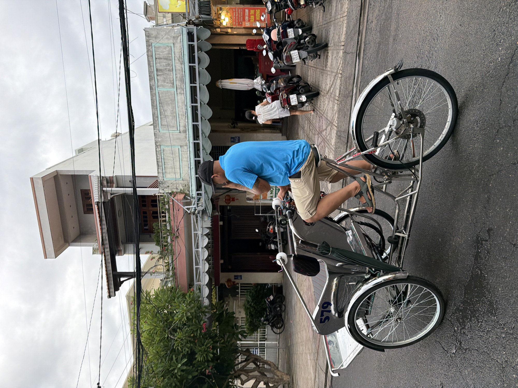 In Nha Trang, Vietnam, a man wearing a blue shirt and tan shorts rides a three-wheeled bicycle. The bicycle is made of metal and has a silver seat. The man is wearing a black baseball cap. In the background, a woman in a white shirt and shorts walks away from a row of parked motorbikes. The buildings in the background are two-story structures with balconies. The street is paved with asphalt, and there are power lines overhead. The sky is overcast.