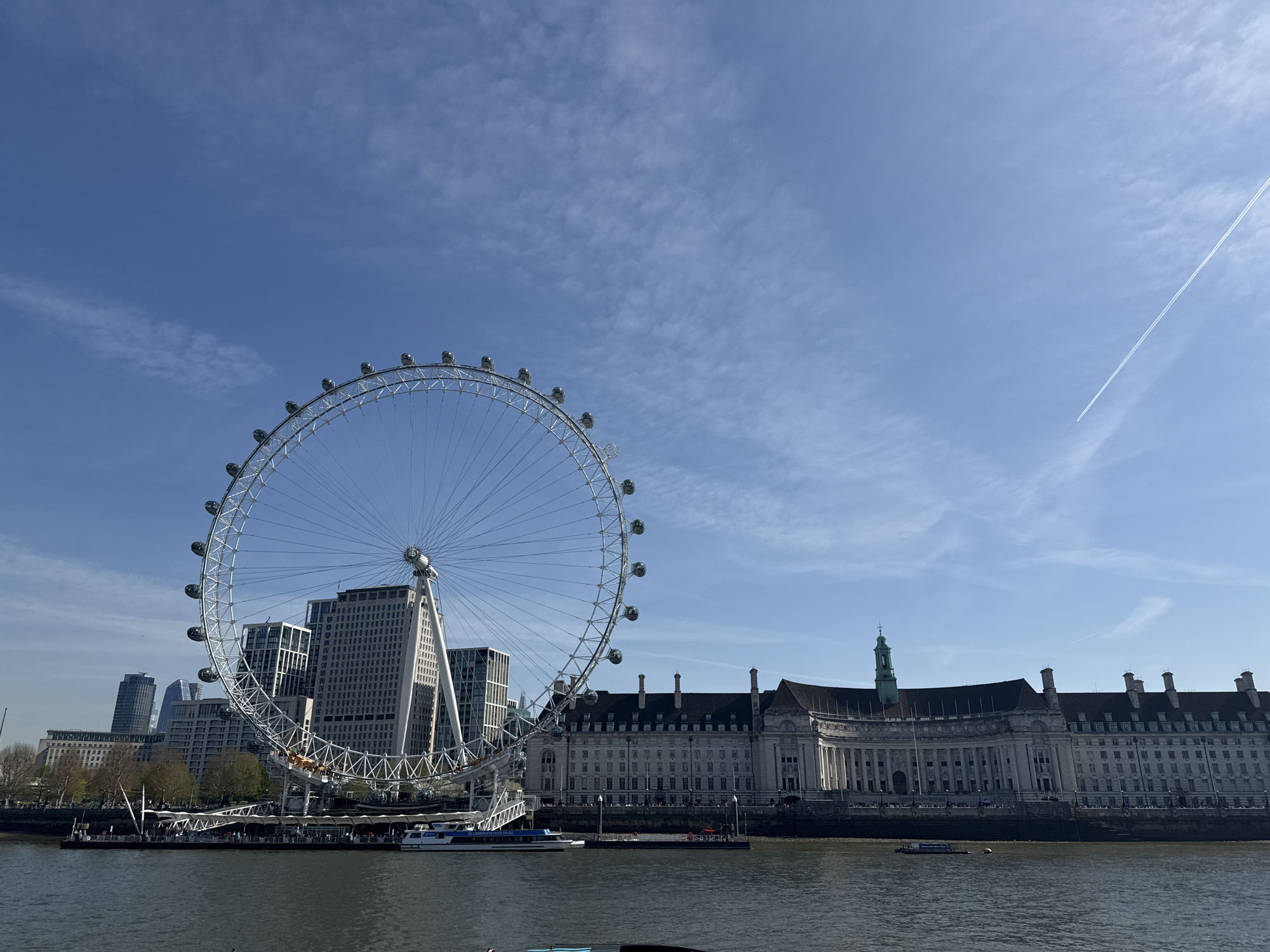 The image shows the London Eye, a large Ferris wheel on the bank of the River Thames in London, United Kingdom. The Houses of Parliament are visible on the opposite bank of the river. Several boats are on the river. The sky is blue with some scattered clouds and a contrail from an airplane.