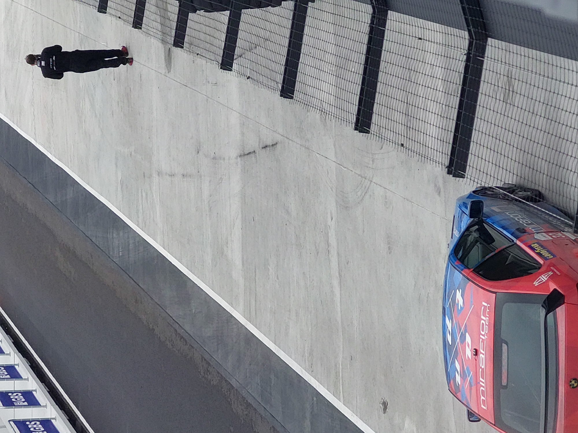 The image presents an elevated, angled view of a motor racing track. A race car, predominantly red with blue accents and markings including the word "mirafiori team," is parked on the track’s surface near a wire fence. Tire tracks are visible on the concrete. A person dressed in dark clothing, possibly wearing red shoes, is lying down on the track surface near the fence, seemingly still. The location is noted as being in the city of Unknown, Unknown.