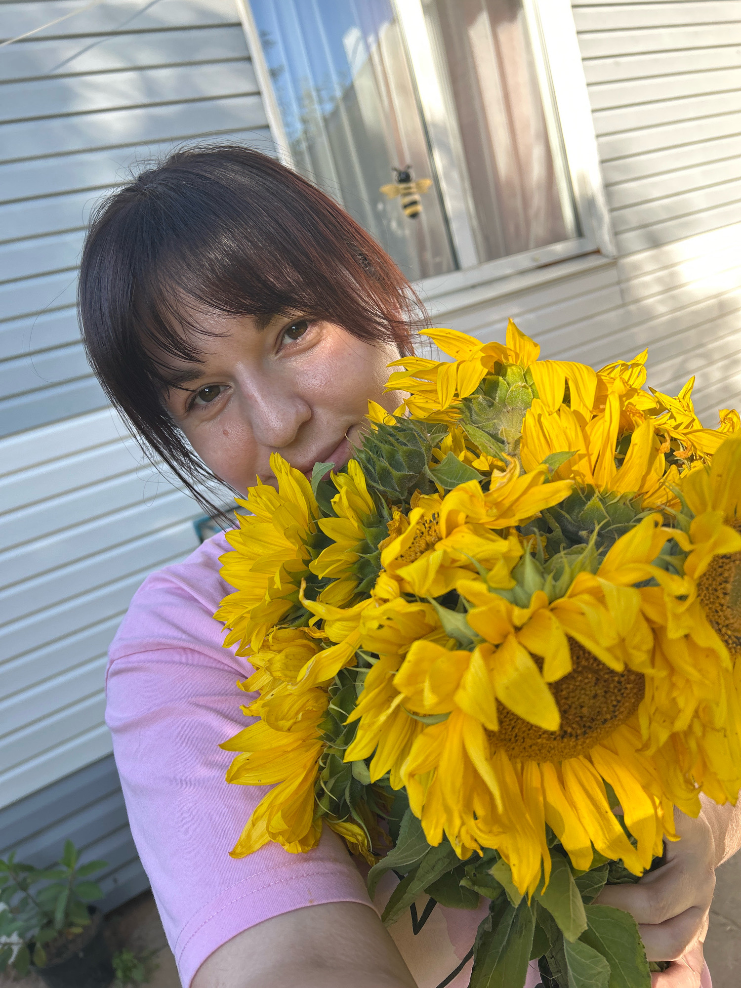 A young woman with dark hair and bangs is holding a large bouquet of sunflowers. She is wearing a pink t-shirt. The background appears to be the exterior wall of a building, possibly a house, with horizontal siding. There is a window in the background with a reflection that appears to show a small bee or insect decoration. The image was taken outdoors in daylight, as indicated by the shadows and bright lighting. The setting is implied to be a residential area.