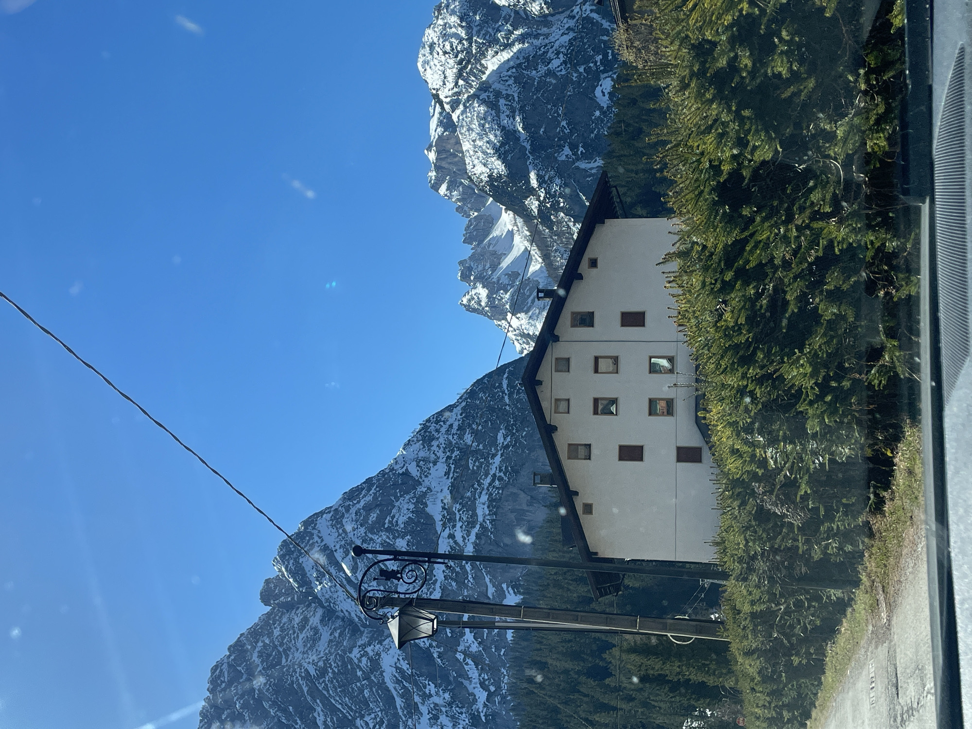 A daytime shot of a light-colored building, possibly a lodge or hotel, situated in a mountainous region, likely the city of Sappada/Plodn/Sapade, Italy. The building is multi-storied with numerous windows. It is flanked by evergreen trees and snow-capped mountains in the background under a clear blue sky. A power line and street lamp are visible in the foreground, suggestive of human presence. The photo is taken from inside a vehicle.