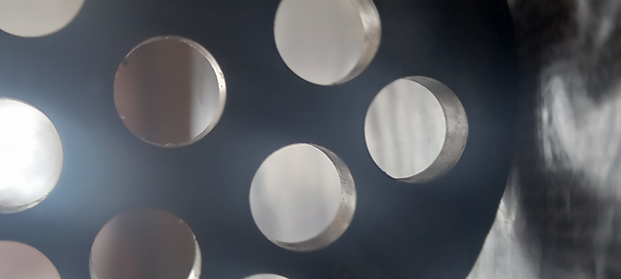 The image shows a close-up of a dark gray metal plate with several circular holes. The holes appear to have reflective surfaces inside them, giving the impression of metal discs embedded in the plate. The lighting is uneven, with some areas of the plate brighter than others. The right side of the image is blurred and out of focus. There is no evidence of the location being Kryvyi Rih, Ukraine, nor of any people or interactions.