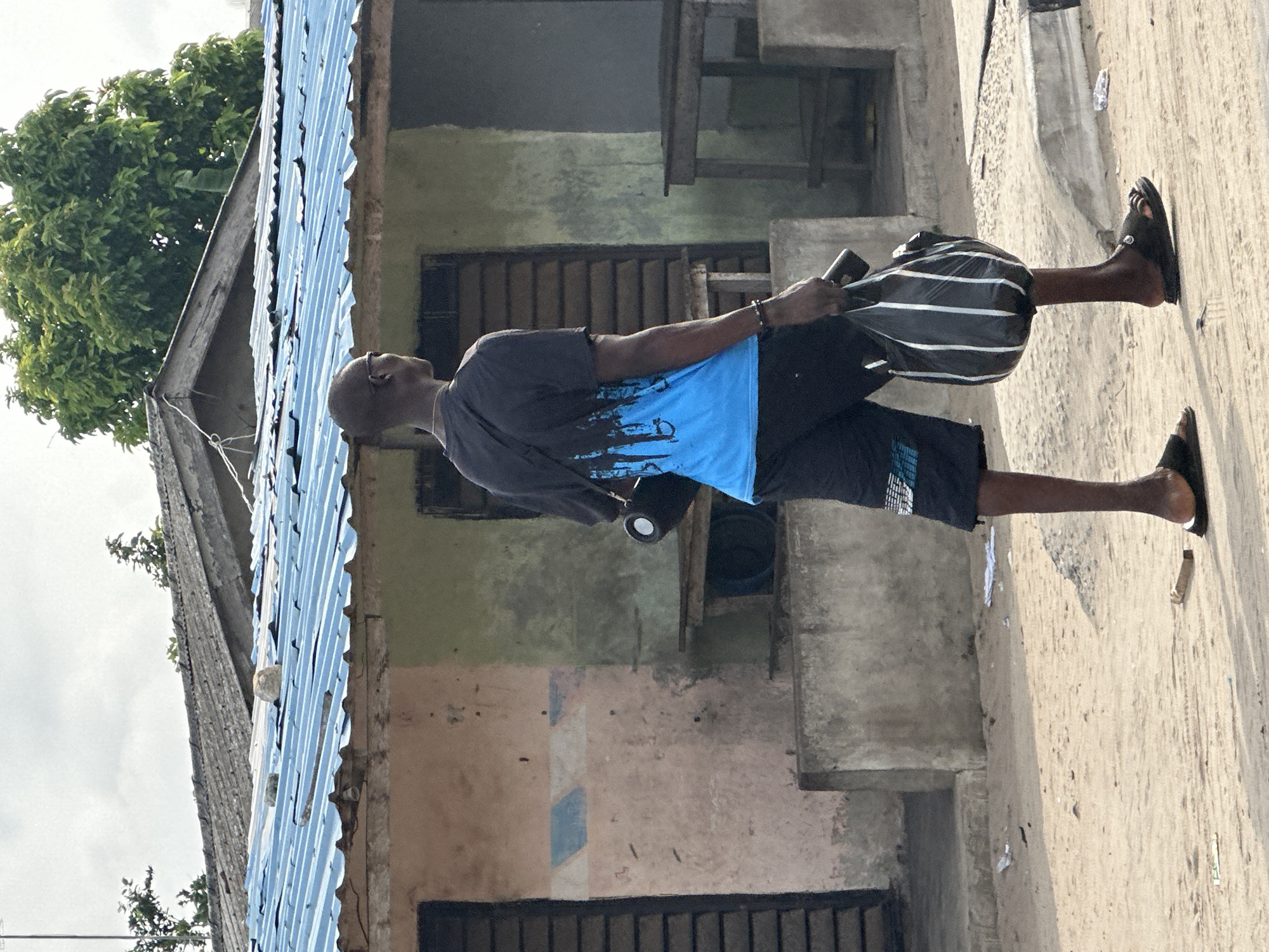 In Badagry, Nigeria, a Black man walks on a dirt path in front of a building with a blue corrugated metal roof. He is wearing a dark t-shirt with a blue and black design on the back, black shorts, and black sandals. He is carrying a black and white striped bag in his right hand and a black cylindrical object in his left hand. The building has a textured, light-colored exterior with sections of green and pink paint. There are wooden benches or tables visible inside the open doorways of the building. The ground is sandy with some scattered debris. Trees are visible in the background.