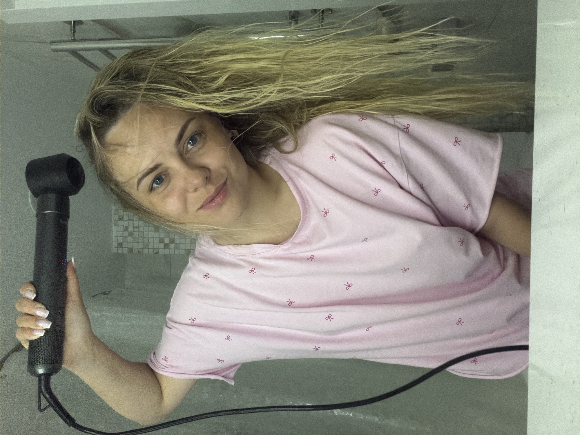 An adult female, estimated to be between 20 and 40 years old, is depicted in a close-up, reflected image within a bathroom setting. She has blonde, wet, and disheveled hair, and blue eyes, with a faint smile directed towards the camera. She is wearing a light pink short-sleeved top adorned with multiple small, dark pink bow-tie patterns. Her right hand, featuring a white-tipped French manicure, is elevated, grasping a black, cylindrical hairdryer equipped with a nozzle attachment. Blue indicator lights are visible on the hairdryer's body, from which a black power cord extends downwards. The background consists of a light-colored tiled wall section on the left, a metallic fixture (likely a towel rack) on the upper right, and a pale horizontal surface in the lower foreground. The scene represents a domestic interior. The image contains no visual evidence to confirm the location as the city of Votkinsk, Russia.