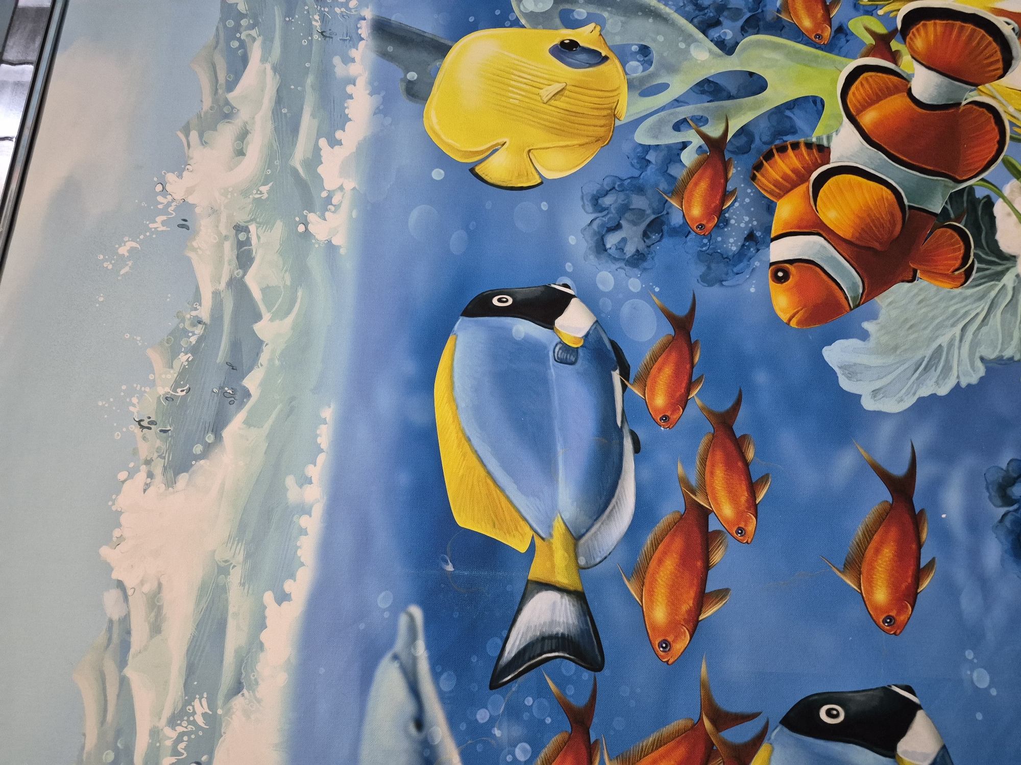 The image depicts a painting of a vibrant underwater scene. Various colorful fish, including clownfish, yellow tangs, and blue tangs, are interspersed among aquatic plants and bubbles against a blue background. On the left side of the image, there's a depiction of what appears to be waves or ocean foam. There are no people or objects besides the painted elements. The location in the city of Tobolsk, Russia, is not directly discernible from the image.