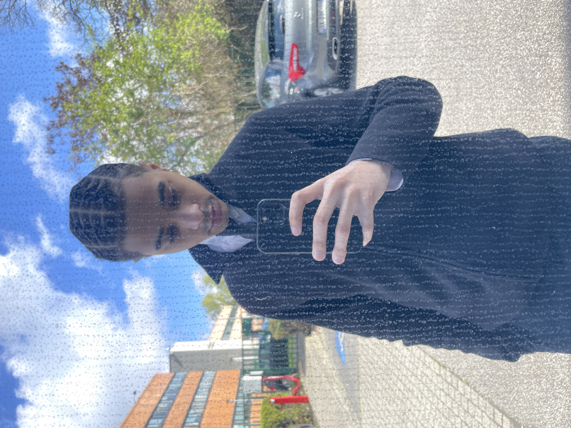 A young male subject, characterized by dark braided hair, is depicted from the upper torso upwards, looking downward at a black smartphone held in his right hand, suggesting a self-portrait or video recording. He wears a dark, collared jacket over a white collared shirt. The image is captured through a reflective surface heavily covered in water droplets, partially obscuring the view.
The background, visible through the reflective surface, portrays an outdoor urban environment under a blue sky with scattered white clouds. To the left, a multi-story building with an orange-brown brick facade and rectangular windows is present. Adjacent to it, two prominent red vertical bollards stand on a paved area, which includes a faint blue square ground marking. Further right, a dark grey sedan, identifiable as a Citroën DS5 based on the taillight design, is parked. Its French registration plate, FJ-930-MM, is partially visible. Green deciduous trees with full foliage are situated in the mid-ground and right side of the frame. This scene is situated in Montigny-le-Bretonneux, France.