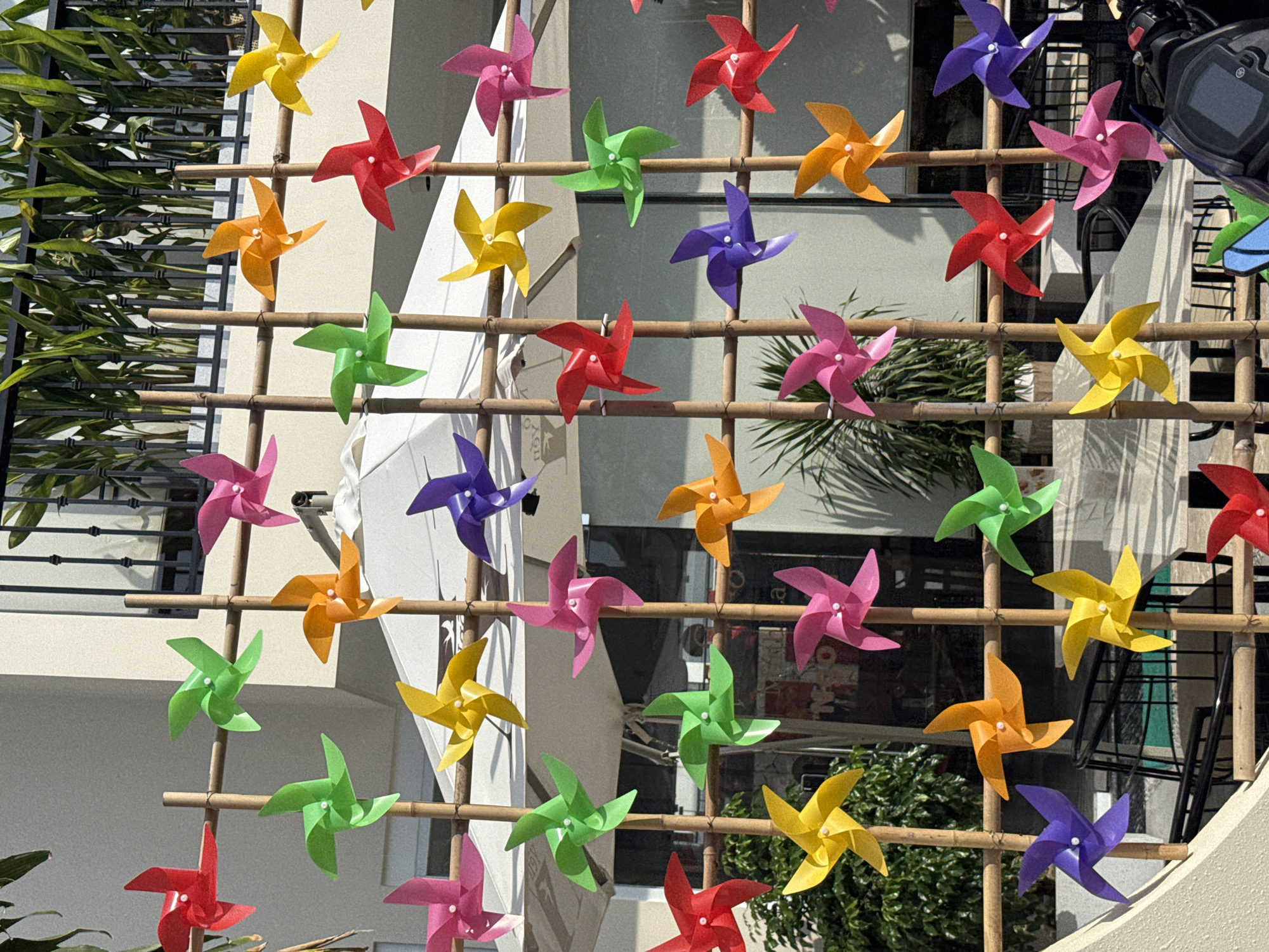 In Nam Nha Trang Ward, Vietnam, a brightly colored arrangement of pinwheels decorates an outdoor setting. The pinwheels, made of red, green, yellow, orange, pink, and purple plastic, are attached to a bamboo frame. They are positioned at varying heights and angles, some spinning in the breeze. Behind the pinwheels, a white umbrella provides shade, and a black metal railing with green foliage is visible. The scene appears to be in a public space, possibly a cafe or restaurant courtyard, with a light-colored building facade in the background. The overall impression is one of a vibrant and cheerful decoration.