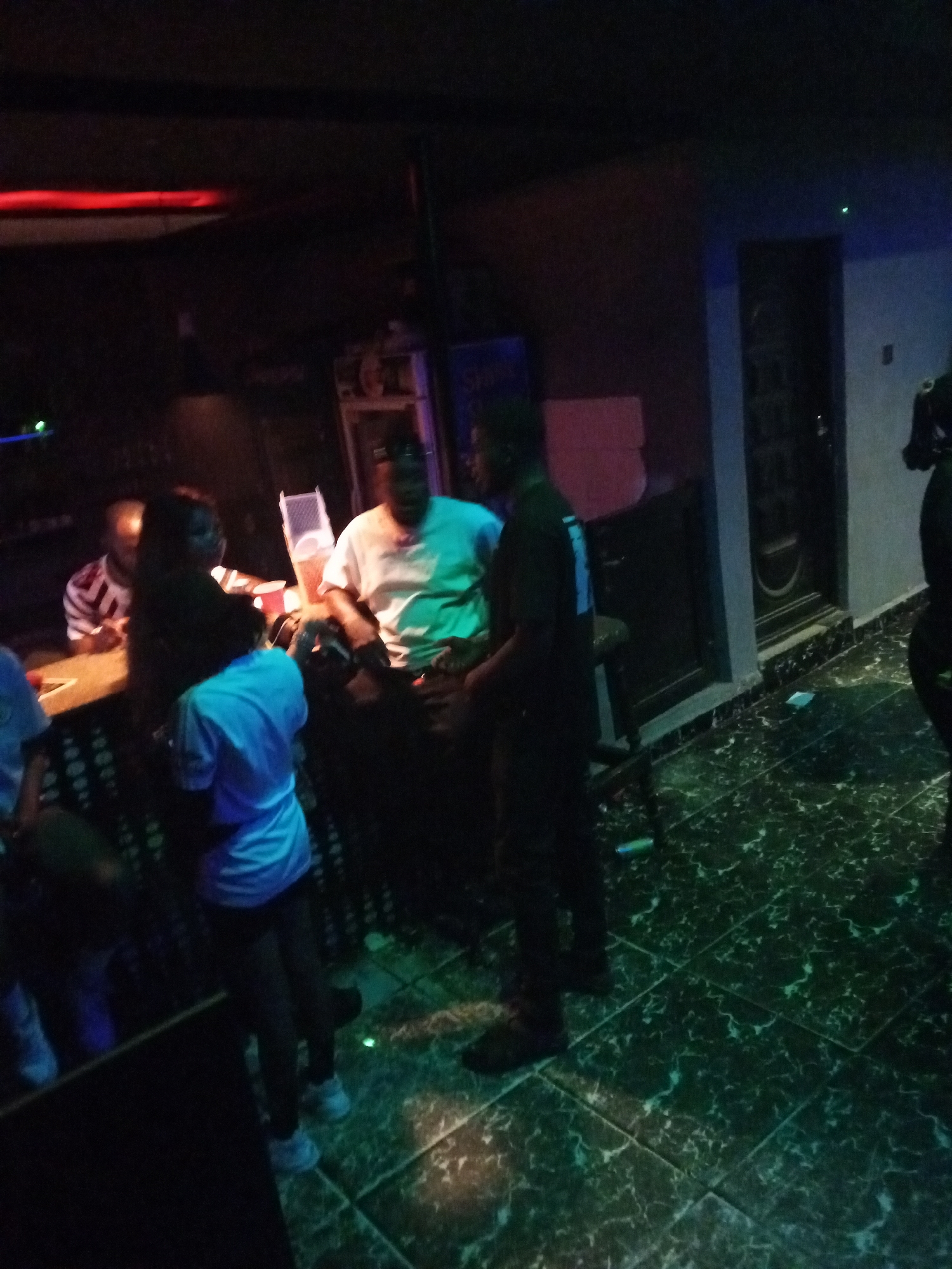 Scene in dimly lit interior of Unknown, Unknown with marble tiled floor. Several individuals are present, including at least one woman wearing a blue shirt who stands facing a bar. The bar has patrons, and one man is seated on a stool. A person dressed in dark clothing is standing near the bar. There is a door visible in the background. Above the bar are red lights.