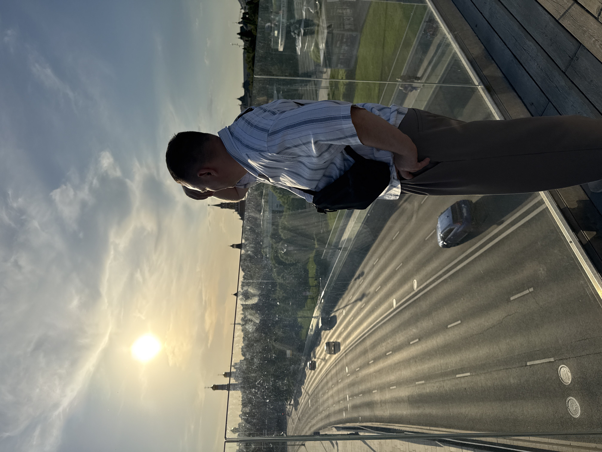 The image depicts a man looking down from a high vantage point, likely a bridge or observation deck, in Moscow, Russia.
**People:**
* A man with short hair, wearing a light-colored striped short-sleeved shirt and brown pants. He has a black bag slung across his body. He is leaning forward, looking down at the scene below.
**Objects:**
* A transparent barrier or glass wall separates the man from the scene below.
* A multi-lane highway is visible below, with cars driving on it.
* Buildings with towers and domes, characteristic of Moscow architecture, are seen in the distance.
* Roadway markings
* Street Lights
**Actions/Interactions:**
* The man is looking down at the road and distant buildings.
**Environment:**
* The image suggests it is either sunrise or sunset, given the low sun and the lighting conditions.
* The weather appears clear.
* The vantage point is high, providing a wide view of the city.
* Green space visible.
**Location:**
* Moscow, Russia, based on the architecture of the buildings in the distance. The vantage point overlooks a major highway in the city.