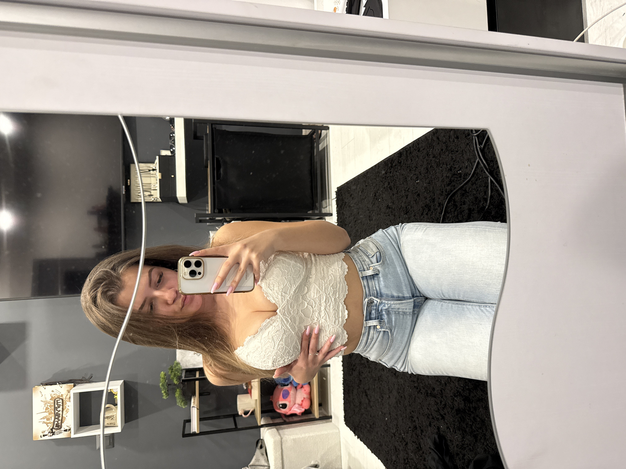 A young woman is taking a selfie in front of a mirror. She is wearing a white lace top and light blue jeans. She is holding her phone in her left hand, and her right hand is partially covering her chest. Her fingernails are painted pink. The background includes a television, a shelf with plants and other objects, a white chair, and a black rug. The location appears to be inside an apartment. There is no evidence to suggest that the location is in the city of Sofia, Bulgaria.