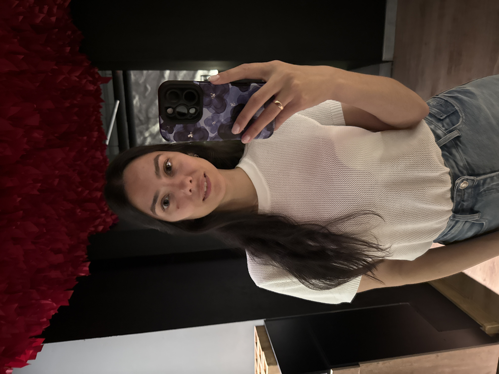 A young woman takes a selfie in an indoor setting. She holds her smartphone, encased in a dark floral case, in her right hand, her wedding ring visible on her ring finger. She is dressed in a white, open-knit, short-sleeved top and blue jeans. Her dark hair falls over her shoulder. Behind her, a wall is adorned with a large, red, textured artwork. Part of a black shelving unit is visible. A television sits on a shelf. The photograph was taken in Chaikovsky, Russia.