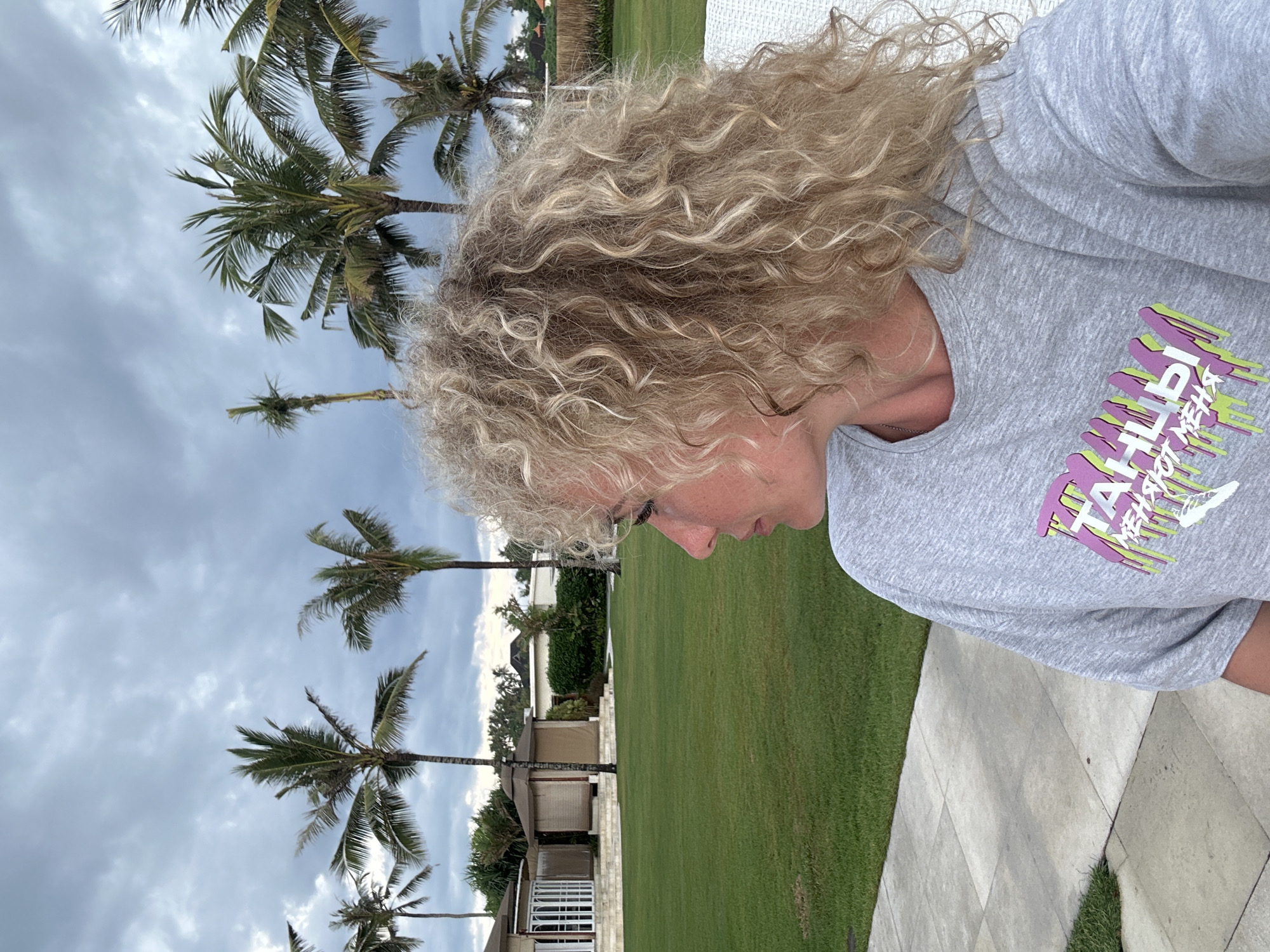 A medium shot, taken from a slightly elevated perspective, features a woman with blonde curly hair, turned slightly to the right, gazing downward. She is wearing a light gray t-shirt adorned with a design in hues of purple and green. The background presents a lush green lawn dotted with numerous palm trees that stretch into the overcast sky. In the distance, a building with a beige roof can be discerned. The photo is taken outdoors in a serene environment. The location is Unknown, Unknown.