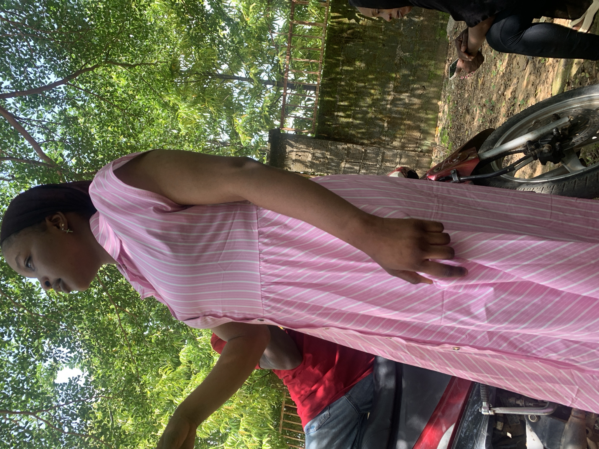 In Akwanga, Nigeria, a person wearing a pink and white striped dress stands near a motorcycle. The scene is outdoors, with lush green trees and foliage providing a natural backdrop. Sunlight filters through the leaves, indicating it is daytime, likely a clear or partly cloudy day. A rough, possibly brick, wall is visible in the background, suggesting an urban or semi-urban environment.
Another person, partially visible and wearing a red shirt and blue jeans, is positioned behind the motorcycle, their arm and shoulder area in view. To the far right, another individual is seen from the waist down, wearing black pants and white shoes, holding a mobile phone. The focus of the image is on the person in the pink dress, with their head tilted down and to the side, appearing thoughtful or perhaps engaged in a conversation. The overall atmosphere seems casual and relaxed, typical of an everyday outdoor setting. There is no visible text in the image.