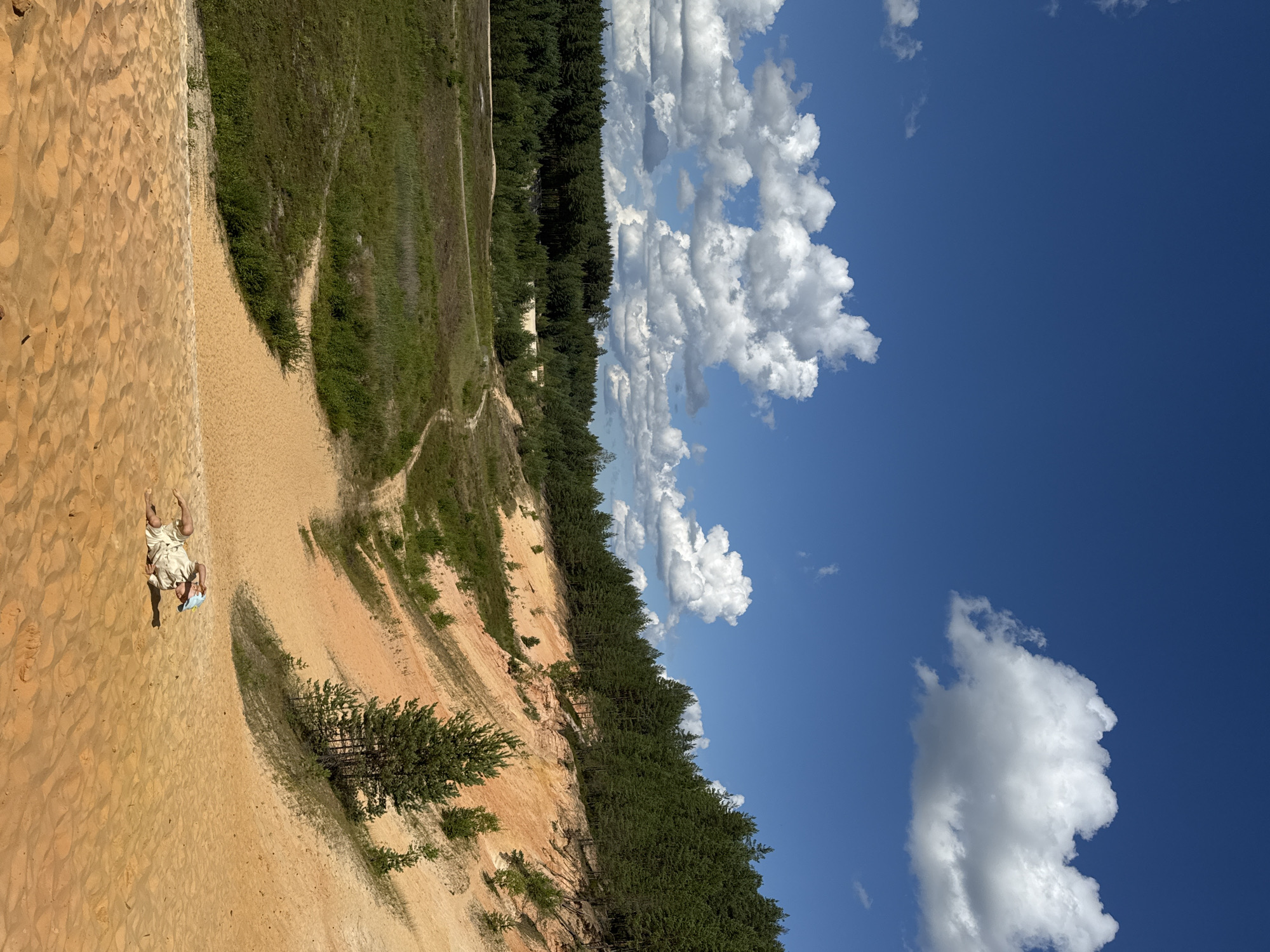 In the Piusa Sand Dunes of Estonia, under a vibrant blue sky dotted with fluffy white clouds, a person is seen lying on a sandy slope. The individual, wearing light-colored clothing and a blue hat, appears to be relaxing or possibly attempting to roll down the dune. The landscape is characterized by rolling sand dunes with patches of sparse vegetation, leading up to a dense forest of pine trees that forms a backdrop. A winding path is visible through the grassy areas, suggesting a natural trail through the terrain. The overall scene depicts a sunny day in a remote, natural environment.
