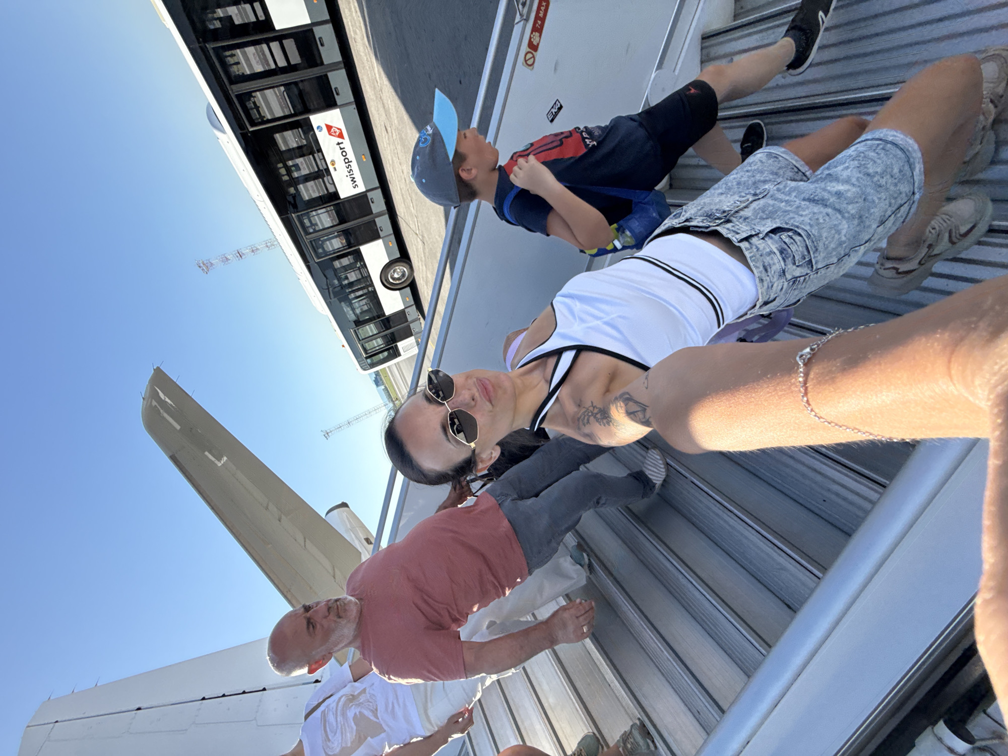In Sofia, Bulgaria, on a sunny day at the airport, a group of people is descending an aircraft's mobile staircase. In the foreground, a woman is taking a selfie with her arm extended, wearing sunglasses, a white tank top, and acid-washed denim shorts. To her left, a man with a bald head and a pink shirt is walking down the stairs. Behind them, another person in a white shirt and light-colored pants is also on the stairs. To the right of the woman, a young boy wearing a baseball cap and a dark t-shirt with a blue backpack is descending the stairs. In the background, a white airport bus with "swissport" branding is parked on the tarmac under a clear blue sky. The environment appears to be an airport tarmac, indicated by the presence of the aircraft stairs, the bus, and the open sky.