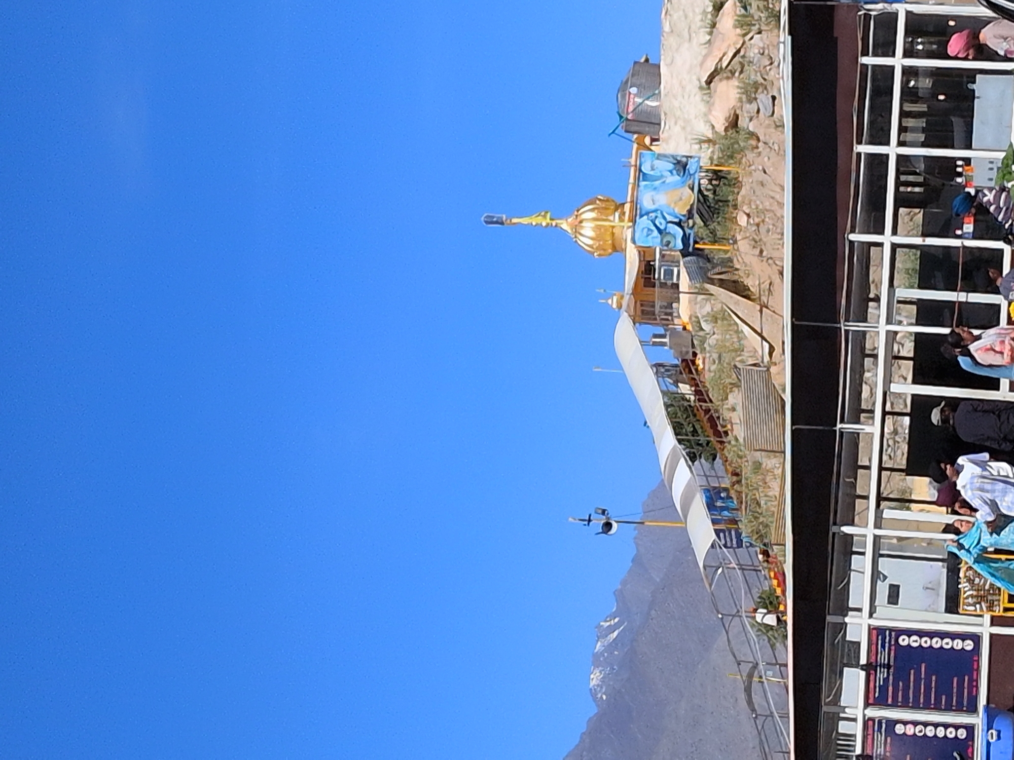 In the city of Chumkar, India, a golden-domed structure with a flag stands atop a rocky hill under a clear blue sky. A white, arched canopy shelters a pathway leading up the hill, where several people are visible walking. At the base of the hill, a modern building with large glass windows and a dark roof is situated, with people gathered in front of it. Signs with text and bullet points are displayed on the left side of the building. In the background, a snow-capped mountain range rises against the horizon.