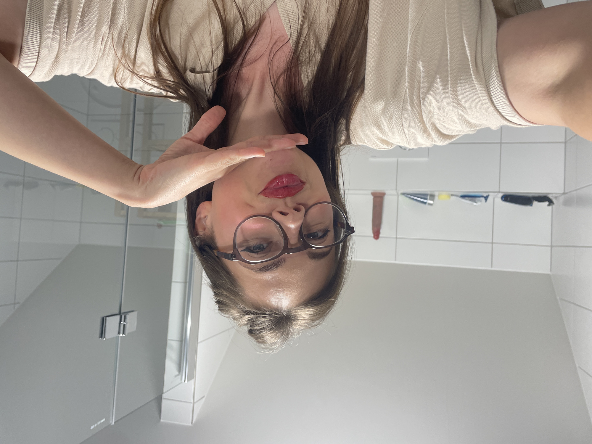 A young woman with brown hair pulled up into a bun, wearing glasses and a beige shirt, is taking a selfie in a bathroom. She is wearing red lipstick and has her right hand raised to her chin. The bathroom has white tiled walls and a glass shower door. On a shelf in the background, there are various toiletries. The image is taken from a selfie perspective, with the woman looking directly at the camera. The lighting suggests an indoor setting, possibly a domestic bathroom. No specific landmarks or indications of Vrchlabí, Czechia are present in the image.