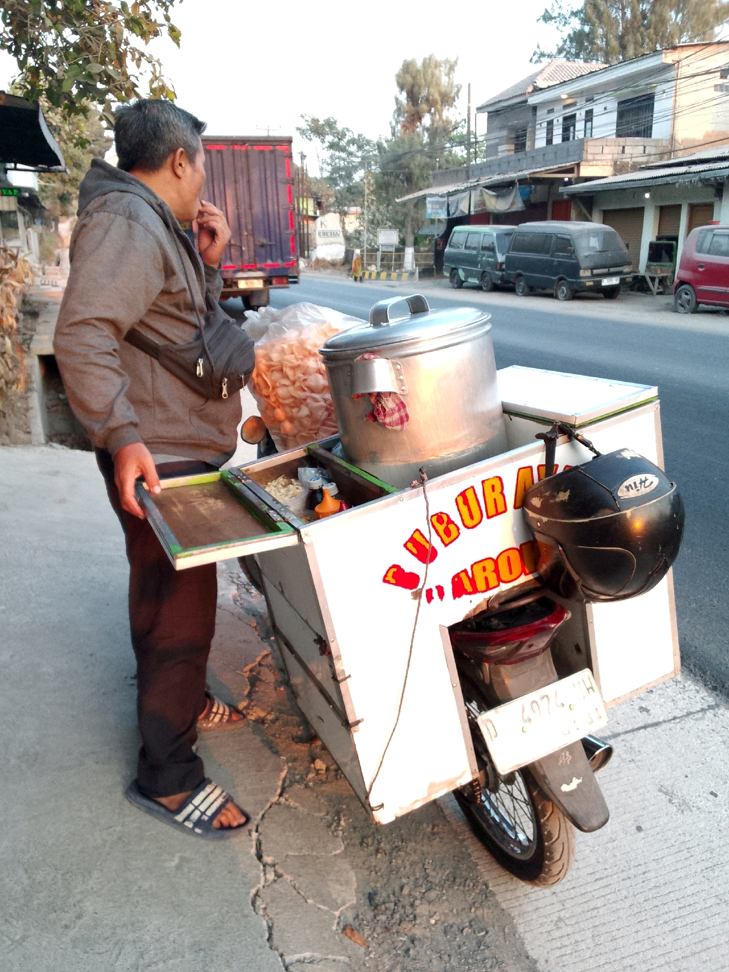 A man in a grey hooded sweatshirt and dark pants stands on a sidewalk next to a motorcycle cart on a street in Gunungmasigit, Indonesia. The man is facing left and has his right hand up to his mouth. He is wearing a dark bag across his chest. The motorcycle cart is white with red lettering that reads "BUBUR AYAM". A large pot sits on top of the cart, and a black helmet is attached to the side. In the background, there are several vans and a red car parked along the street. Buildings with awnings and trees line the street. The sky is overcast.