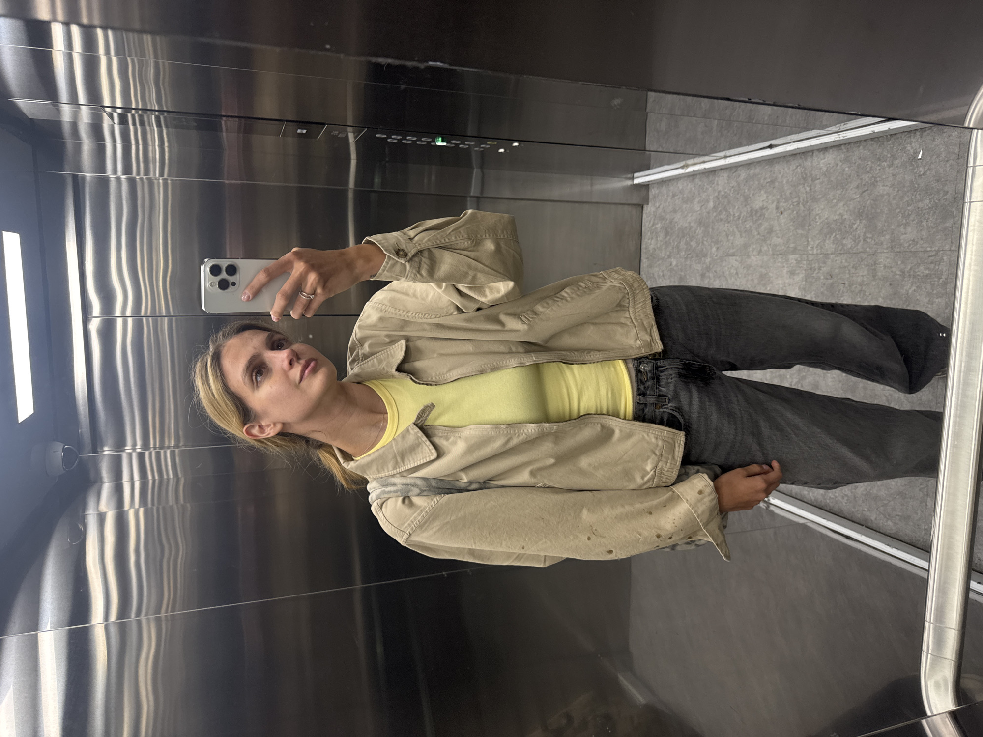A woman is taking a mirror selfie in an elevator. The elevator has stainless steel walls and a grey tiled floor. The woman is wearing a yellow t-shirt, a tan jacket, and dark-wash jeans. She is holding an iPhone in her right hand, and her ring is visible on her ring finger. She has blonde hair that is pulled back. The elevator has a panel of buttons near the top. A security camera is visible in the upper left corner of the elevator.