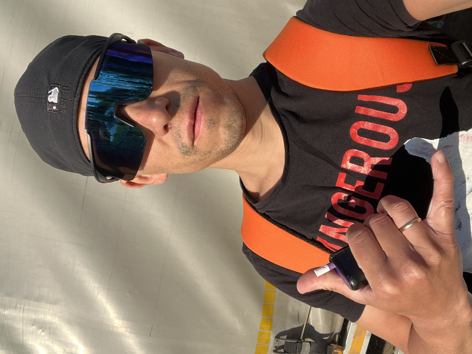 A person wearing sunglasses, a black baseball cap with an MLB logo, and a black t-shirt with large red lettering partially visible, is holding a small black device with a purple accent in their hand. An orange strap goes over their shoulder. The person is wearing a wedding ring. The location, Chaikovsky, Russia, is not discernible from the image.