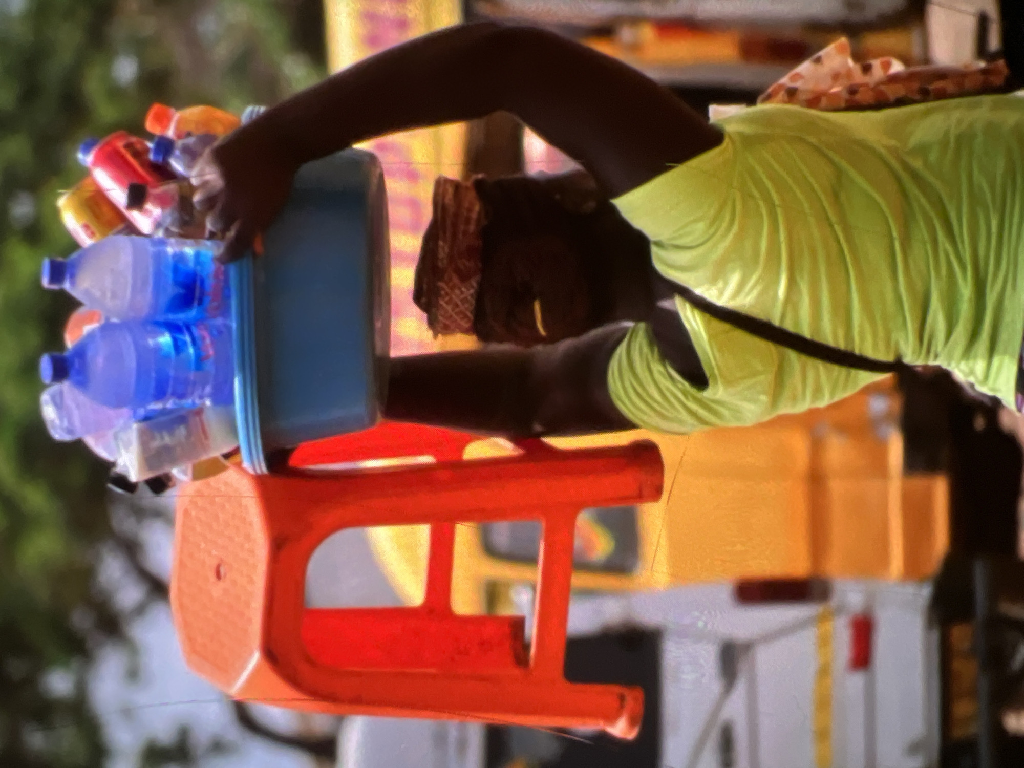 A person is carrying a blue tub filled with bottles of water and other beverages on their head, with a red plastic stool beside them. The person is wearing a bright green shirt and a patterned headwrap. In the background, there are vehicles, suggesting an urban setting, likely a street in Lagos, Nigeria. The scene appears to be a street vendor scene, possibly during the day. The person's left arm is raised, supporting the tub, while their right hand is also near the tub, perhaps adjusting the load. The overall image depicts a busy and vibrant street environment.