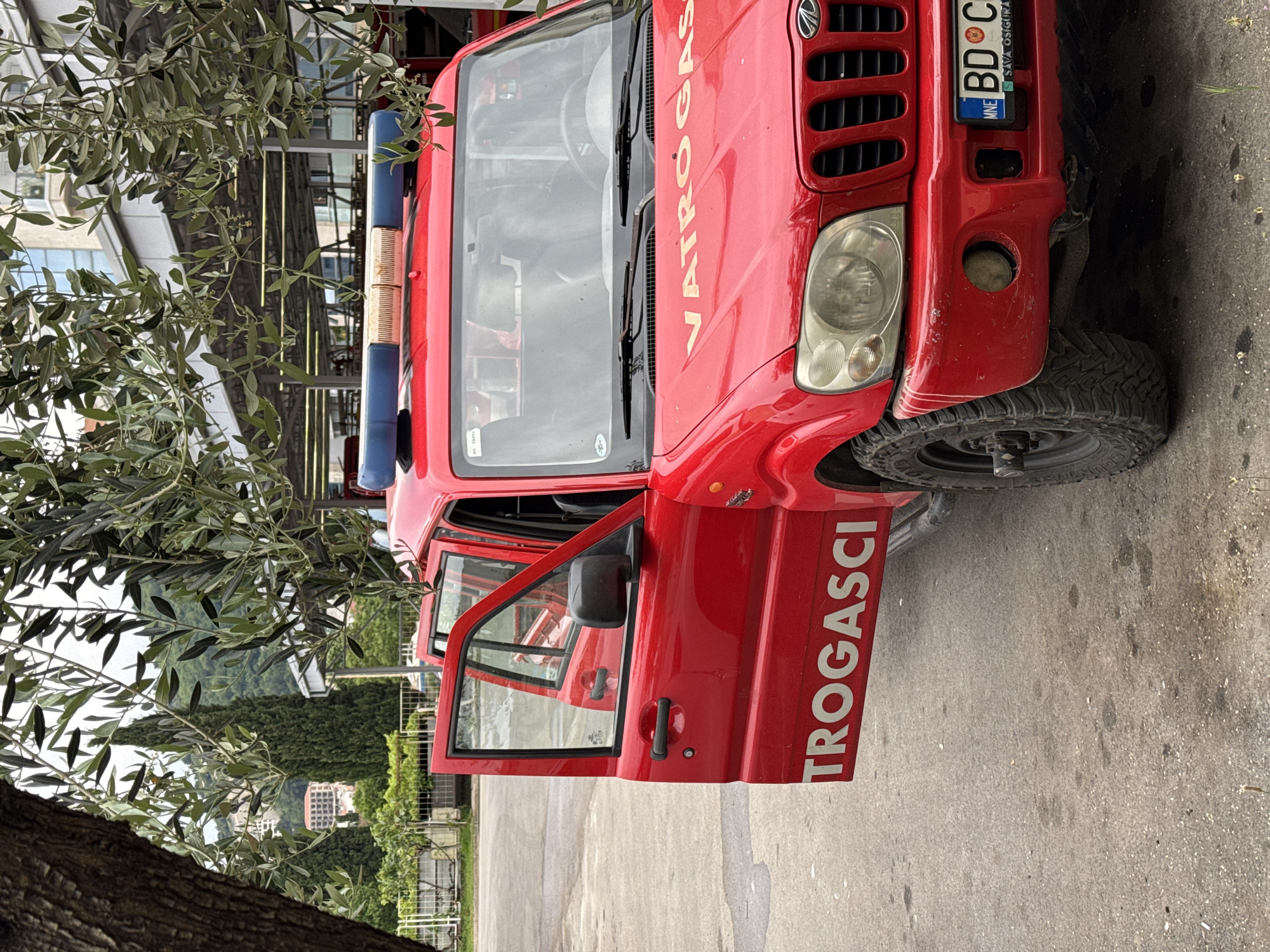 The image shows a red fire truck parked on a street in Budva, Montenegro. The truck has the word "VATROGASCI" printed on the hood and "TROGASCI" on the side. The license plate reads "BD CG." The driver's side door is open. A tree branch with green leaves is visible in the foreground. In the background, there are buildings and greenery, typical of the urban landscape of Budva. The pavement is visible.