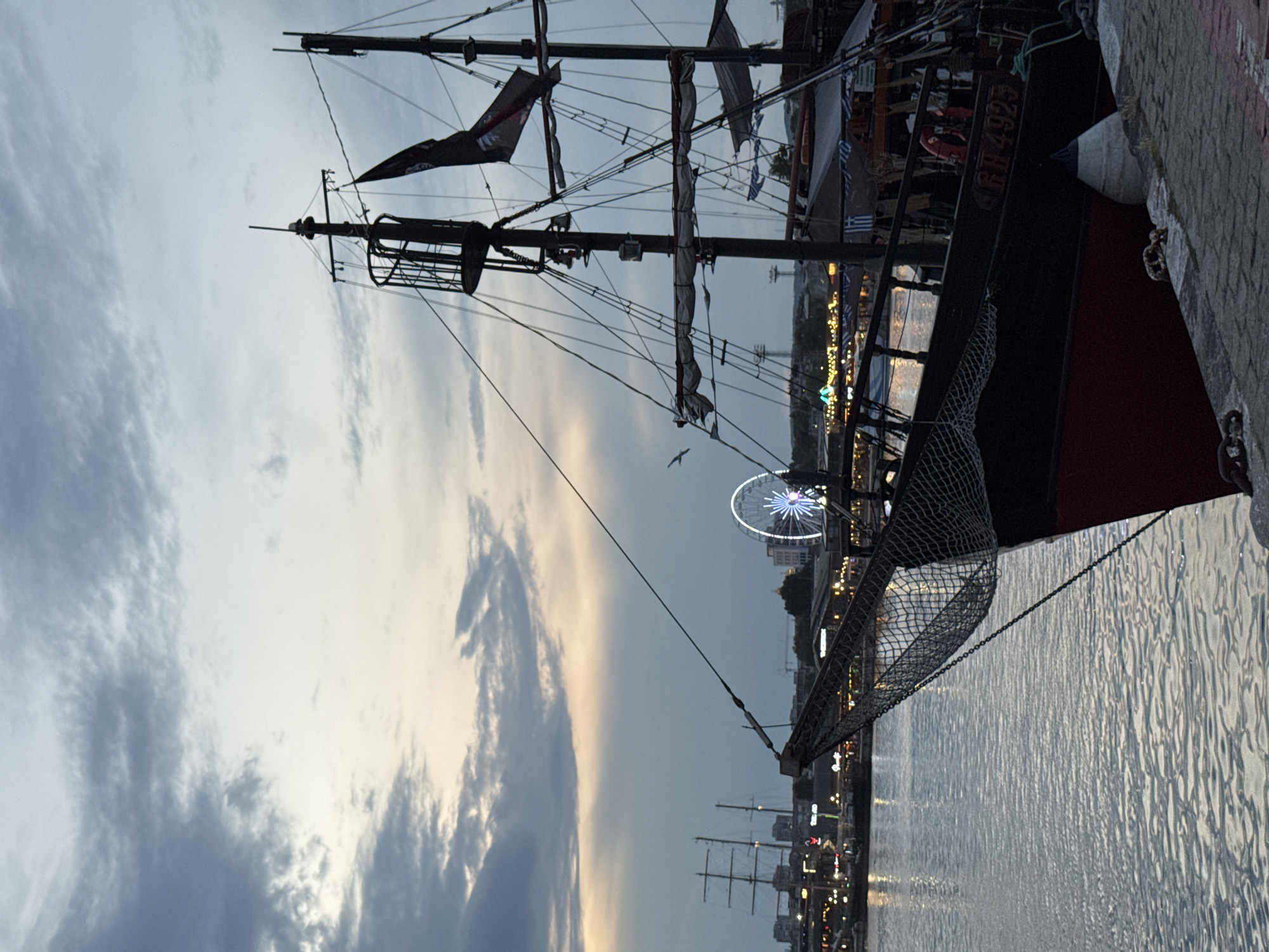 The image shows a harbor scene in Varna, Bulgaria, at dusk. In the foreground, the bow of a dark-hulled pirate-style ship with red trim is docked at a stone quay. A safety net is draped over the bow. The ship has multiple masts, rigging, and a pirate flag. In the mid-ground, the Varna Ferris Wheel is illuminated and prominent, along with other harbor lights and buildings along the waterfront. Further in the distance, another large, multi-masted sailing ship is visible. The sky is cloudy with a soft, diffused light. The water is calm, reflecting the lights and sky. The overall impression is a scenic evening view of a bustling waterfront with historical and recreational elements.
