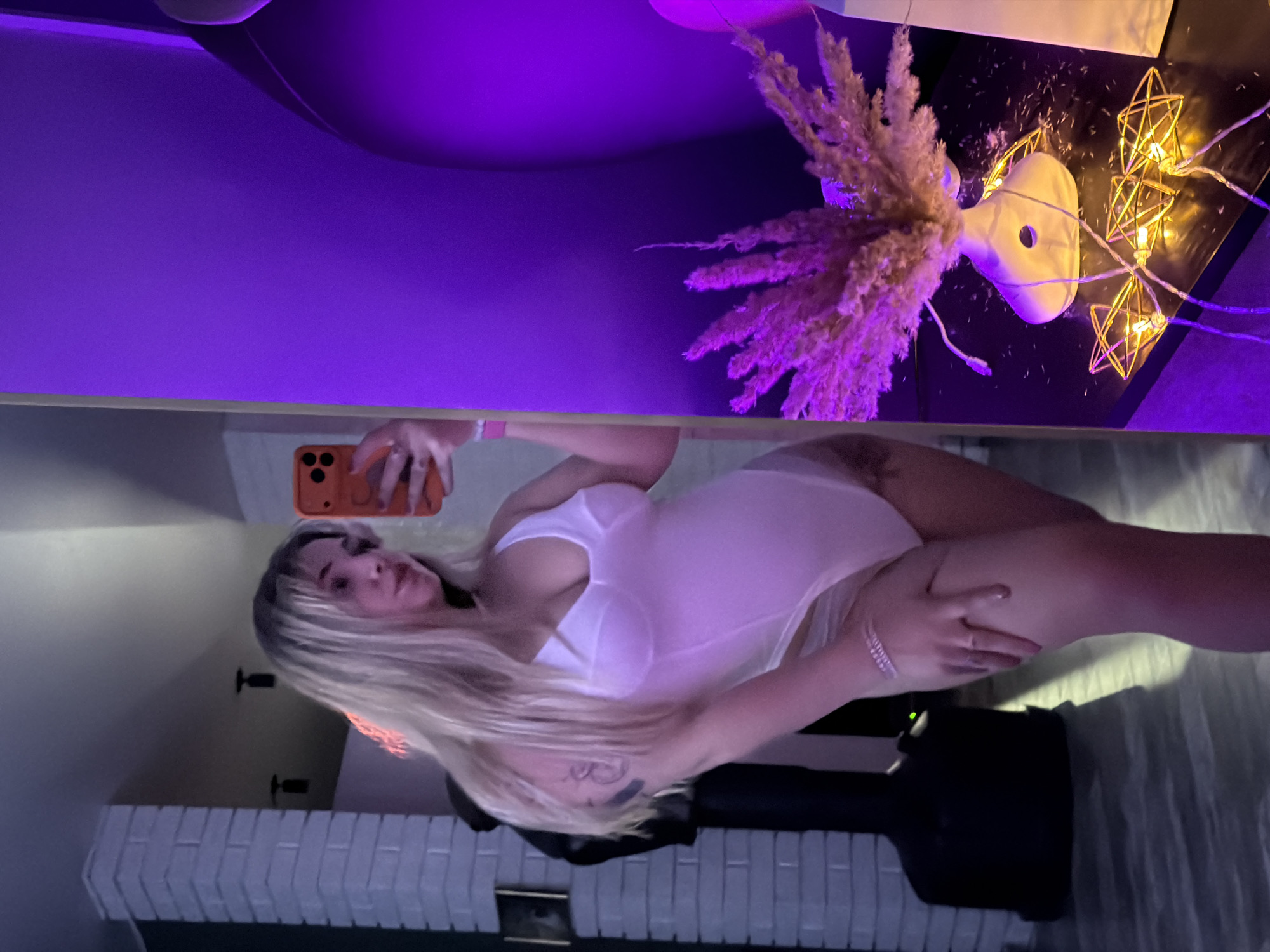 A woman with blonde hair is taking a selfie in a room. She is wearing a white bodysuit. Behind her is a white brick fireplace and a dark object. The room is lit with purple light, and there are decorative items on a table, including a vase with dried flowers and string lights with star-shaped fixtures. The location is indicated as Большая Шильна, Russia, but there is no specific context to verify this within the image.