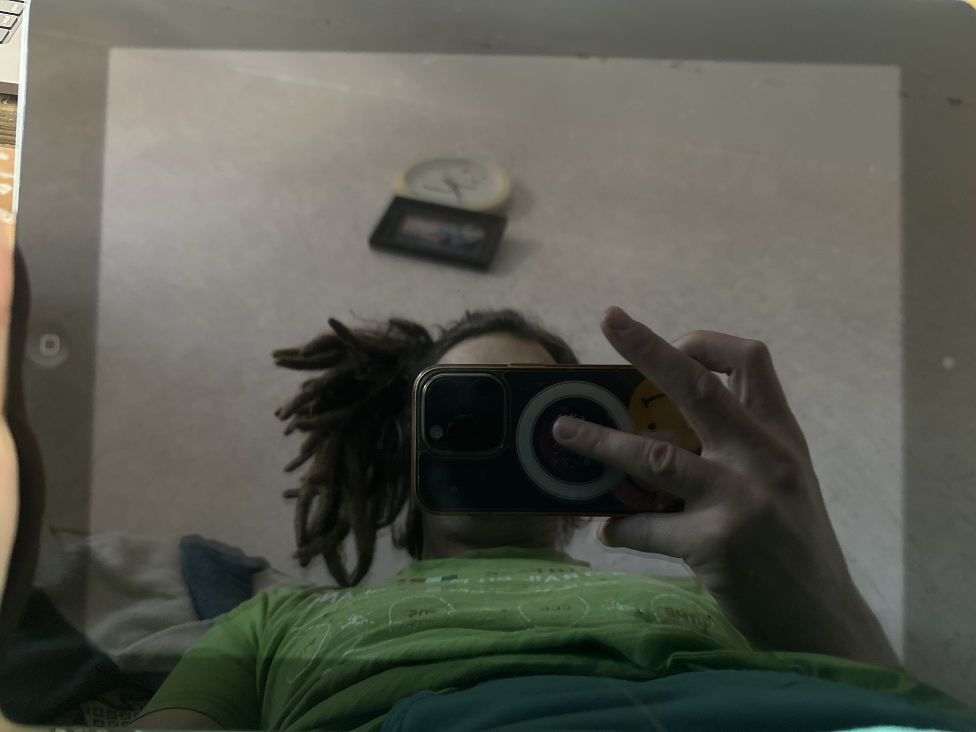 The image is a reflection in a tablet screen, likely of the person holding the phone to take the picture. The person has dreadlocks and is wearing a green shirt. The reflection also captures a blurry background with a clock and a picture frame on the wall. It is impossible to determine if the image was taken in Biysk, Russia or any location without additional information or visual cues specific to the city.
