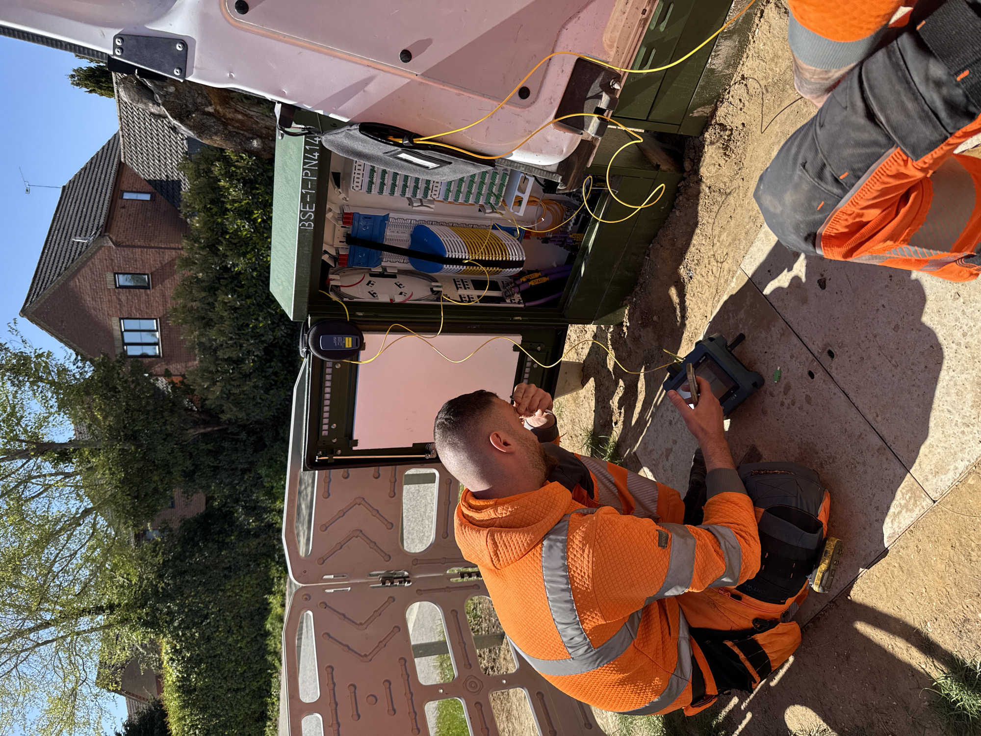 This image captures two workers performing fiber optic network installation or maintenance in a residential area of Mid Suffolk, United Kingdom, under bright, sunny conditions.
In the foreground, a man wearing an orange high-visibility jacket with grey textured panels and matching trousers kneels beside a green, ground-mounted fiber optic cabinet. He is actively working inside the open cabinet, manipulating yellow and other colored fiber optic cables. His left hand holds a small tool, possibly for preparing the fiber, while his right hand operates a handheld electronic device with a screen, likely a fiber optic test instrument like an OTDR (Optical Time-Domain Reflectometer) or a fusion splicer control unit. The device shows a partial "JDSU" or "VIAVI" branding. Another worker, also in orange high-visibility clothing, is partially visible to the right.
The green cabinet contains neatly organized fiber optic cables, including spools of white, blue, and yellow fibers, and various connection points. A yellow fiber optic cable snakes out of the cabinet and across the ground. Text visible on the side of the cabinet reads "BSE-1-PN414". The bright sunlight casts strong shadows on the dirt and paving slabs around the workers.
In the background, a white van, likely a utility vehicle, is partially visible. Beyond that, a brick residential house with a dark tiled roof and multiple windows stands amidst green foliage, including bushes and trees, under a clear blue sky. The overall scene suggests a routine but intricate technical operation to expand or maintain broadband infrastructure.