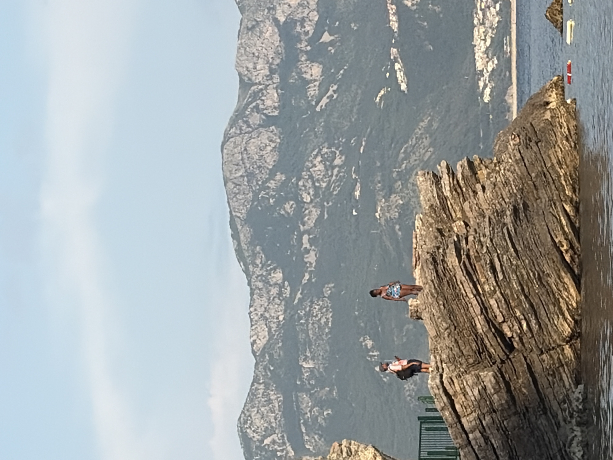 The image depicts a coastal scene in Budva, Montenegro, featuring a rocky outcropping where two individuals are standing. One person, wearing swimwear, is closer to the edge, appearing ready to jump into the water. The other is further back, wearing dark shorts and an orange top. The rock formation exhibits layered, geological stratification.
In the background, a mountainous landscape rises, and a shoreline with some building is visible in the distance. The sea is adjacent to the rock formation. The sky is light blue.
