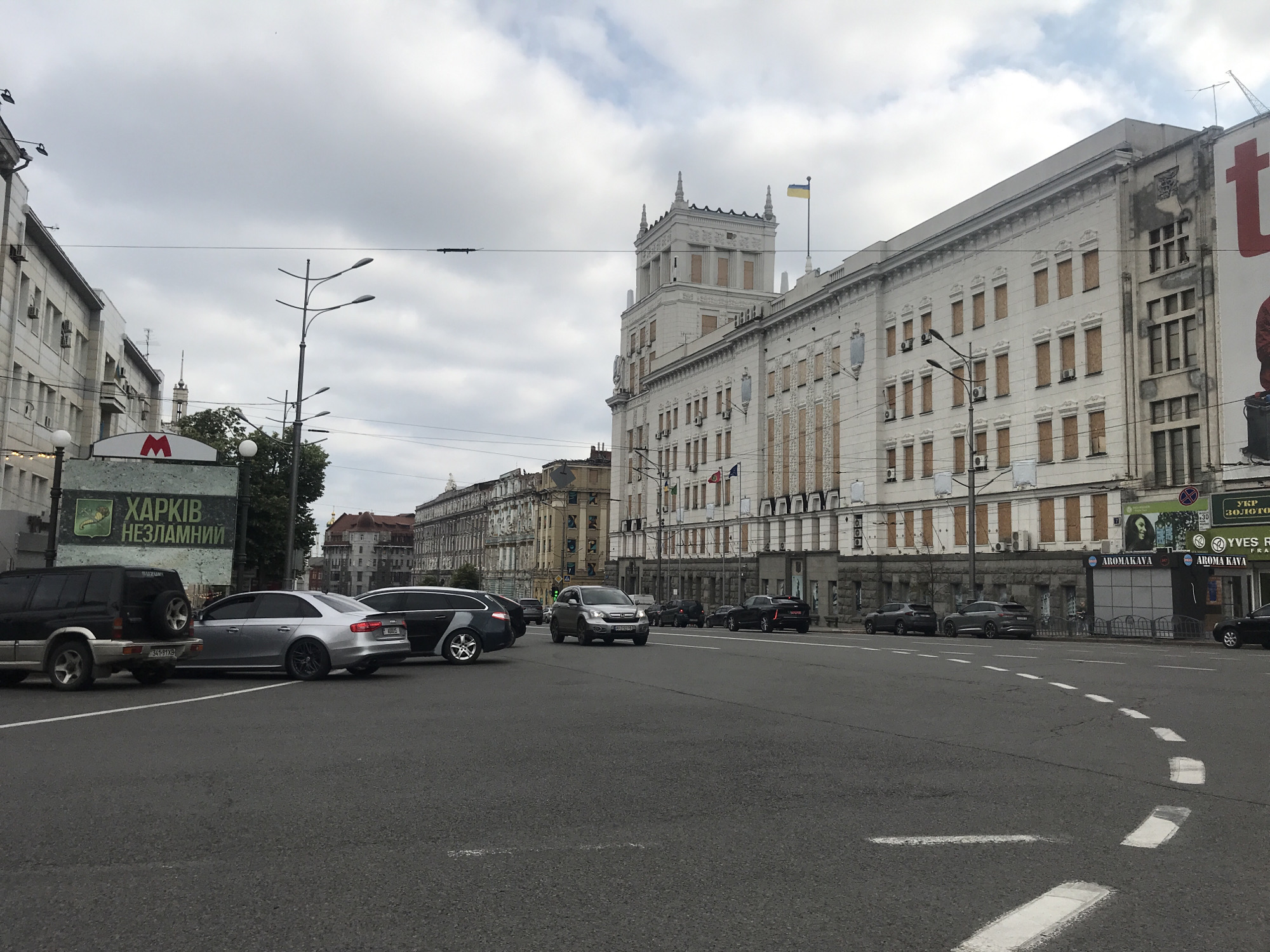 This is an eye-level, medium shot of a city street with multiple lanes of asphalt road. On the left side of the road, a large, light-colored building with boarded-up windows dominates the background. A Ukrainian flag flies from the top of this building. A green billboard with white text is visible to the left of the building. Several cars are parked and driving on the street, including a black SUV, a silver sedan, and a black station wagon. In the background, more buildings line the street, some with architectural details and others appearing older or under construction. Overhead, a cloudy sky suggests overcast weather. There are no visible people in the image, and the scene appears to be a street in an urban environment.