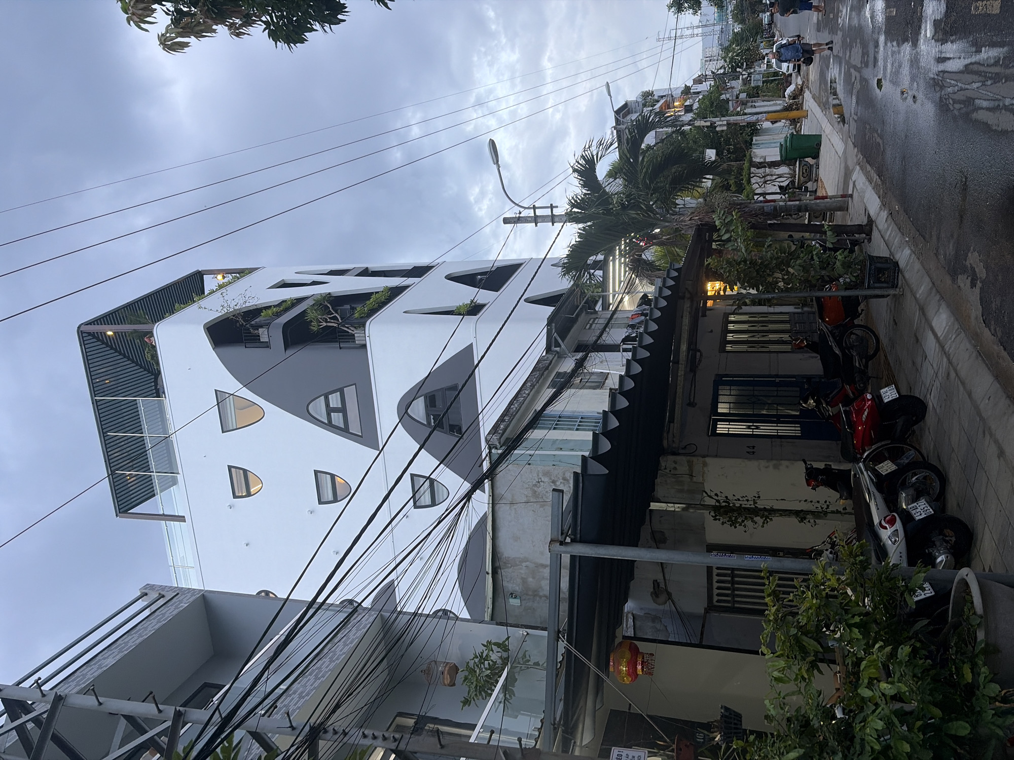 The image depicts a street scene in Đà Nẵng, Vietnam, likely in the early evening or on an overcast day, given the subdued lighting. A modern, multi-story building with a unique architectural design featuring curved, white facade elements and dark grey accents stands prominently on the left side of the street. This building has distinctive arched windows and balconies adorned with greenery.
In the foreground, a street lined with parked motorcycles is visible. These motorcycles are of various colors, including red and white. A mature, leafy plant in a large pot is situated on the left side of the street, obscuring some of the lower portion of the buildings. Several overhead electrical wires crisscross the sky, a common sight in Vietnamese urban environments.
Further down the street, the architecture transitions to more traditional buildings, some with corrugated metal awnings extending over the sidewalk. There are also a couple of streetlights visible, one with a modern design and another, older style. A few individuals are walking on the wet pavement further down the street, suggesting recent rain. One person, a man in a blue shirt and dark shorts, is carrying a dark bag. Another individual in a light blue shirt and darker shorts is walking away from the camera. Two green trash bins are placed along the sidewalk. The road surface appears wet and reflective, indicating moisture. The overall impression is of a typical urban street in a Southeast Asian city, with a blend of contemporary and older structures.