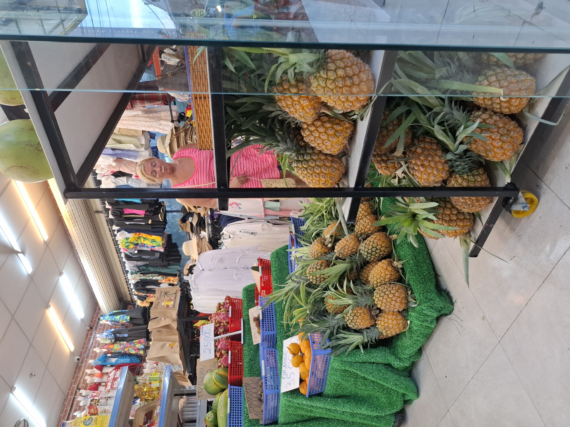 The image depicts the interior of a small shop located in Bac Nha Trang Ward, Vietnam. The shop appears to be a fruit stand or a market stall that also sells clothing. In the foreground, an assortment of pineapples are displayed on shelves, with some also resting on a green, grass-like mat on the floor. Other fruits, such as oranges and dragon fruit, are visible in blue and red baskets on tiered displays. The background shows racks of clothing, including dresses and shirts, and shelves stocked with various items. A woman in a pink sleeveless top is standing behind a counter, appearing to be attending to the shop. The lighting suggests it is daytime, with artificial lights illuminating the interior. The overall impression is of a bustling local marketplace.