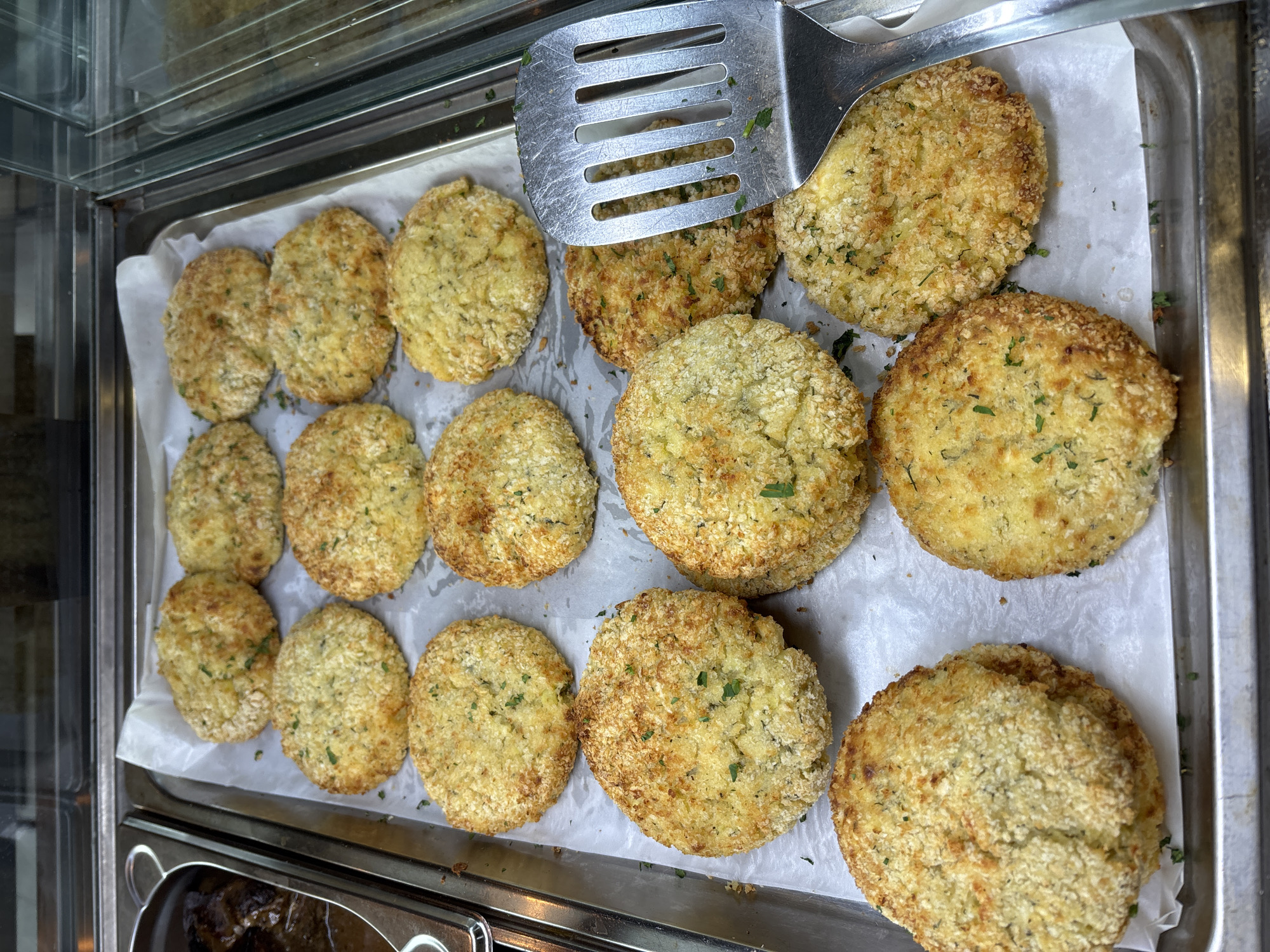 The image shows a metal tray lined with parchment paper, holding about 15 round, breaded and fried fritters or patties, likely vegetarian or vegan based on their color and texture. A metal spatula rests on top of some of the fritters. The tray appears to be inside a refrigerated display case, suggesting a deli or cafeteria setting. There's a glimpse of another food container at the bottom of the frame. Given the user's prompt to include Sofia, Bulgaria, it's implied this scene is located in a food establishment there. No people are visible in the image.
