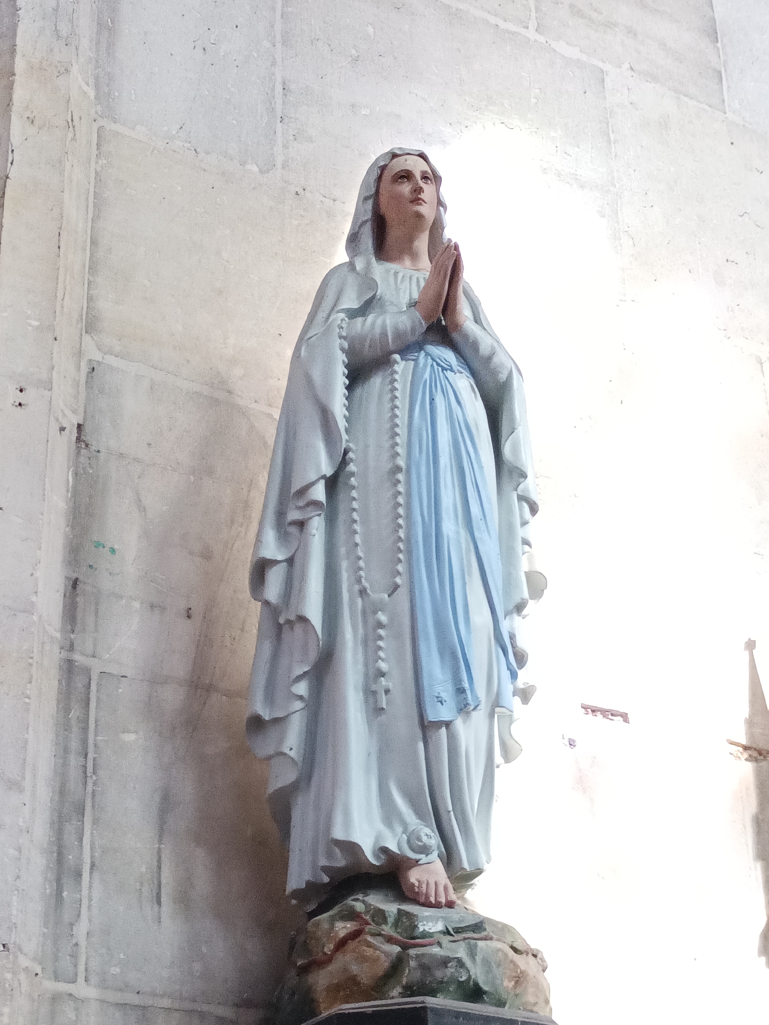A full-body, low-angle, vertical photograph captures a statue of the Virgin Mary standing in a prayerful pose with her hands clasped and eyes looking upwards. She is dressed in a light blue robe with a white mantle draped over her head and shoulders, and a light blue sash cinches her waist. A rosary hangs around her neck, its beads visible against her robe. The statue stands on a rocky base, which is set against a textured, pale stone wall. The wall appears to be part of a church interior, with a bright light source on the right side of the image creating a strong contrast and casting shadows. The statue is centrally located in the frame, with the wall filling the background. This statue is located in the Église Saint-Thomas de Touques in Le Neubourg, France.
