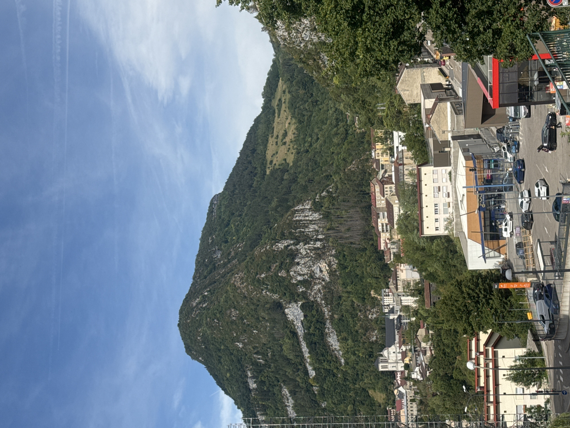 This is a rotated, eye-level, outdoor photograph of a town nestled at the base of a large, verdant mountain. The mountain dominates the left side of the frame, its steep slopes covered in dense green foliage, with lighter patches of rock showing through. A bright blue sky with wispy white clouds occupies the top left quadrant.
To the right of the mountain, the town unfolds, characterized by a mix of buildings with light-colored facades, some featuring red accents and balconies. Residential buildings and a larger white structure with a dome, possibly a church, are visible amongst the trees. A parking lot with several cars is present, and a red-fronted building with large glass windows appears to be a commercial establishment. Some of the cars are black, white, and blue.
The scene suggests a daytime setting, likely late morning or early afternoon, given the bright sunlight casting shadows. The weather appears clear and sunny. The presence of parked cars and the general layout of the town indicate a populated area. No specific activity or event is discernable, other than general town life. There is an orange sign with white text visible in the parking area.
The location is Saint-Claude, France.